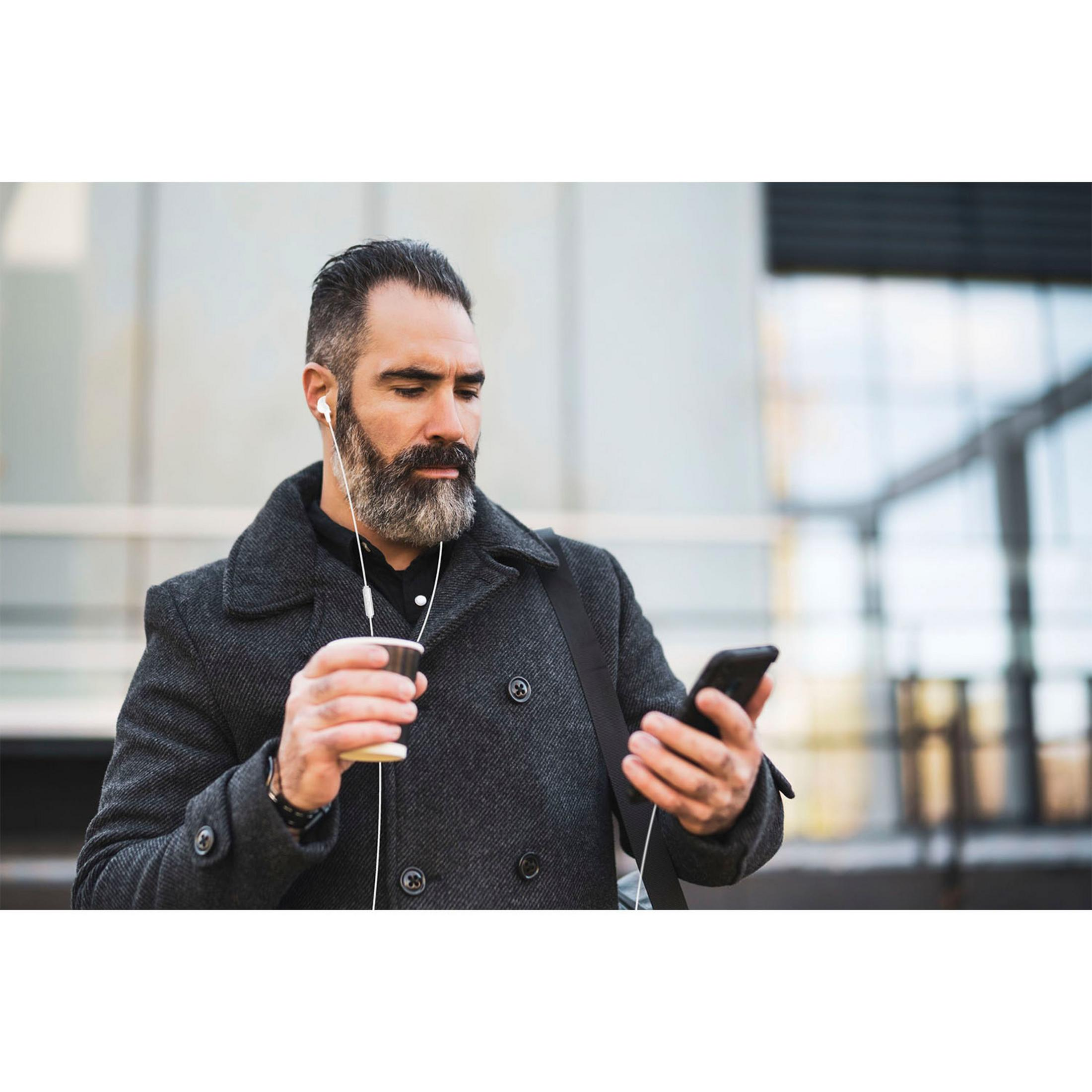 A man with a beard listens to music on earbuds, holding a phone and a cup.