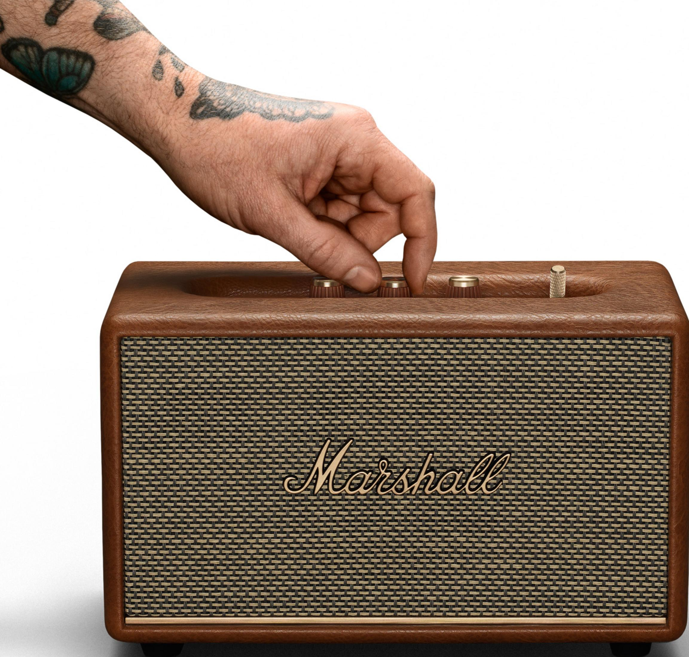 Una mano tatuada ajusta los mandos de un amplificador Marshall marrón. Fondo blanco.