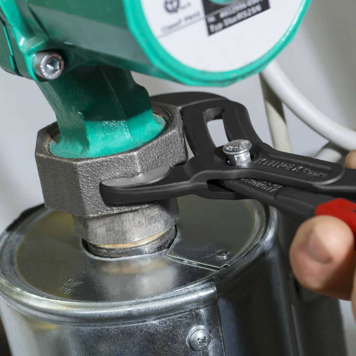 Eine Zange greift eine Mutter an einem grünen Pumpengehäuse, das mit einem Metallbehälter verbunden ist.