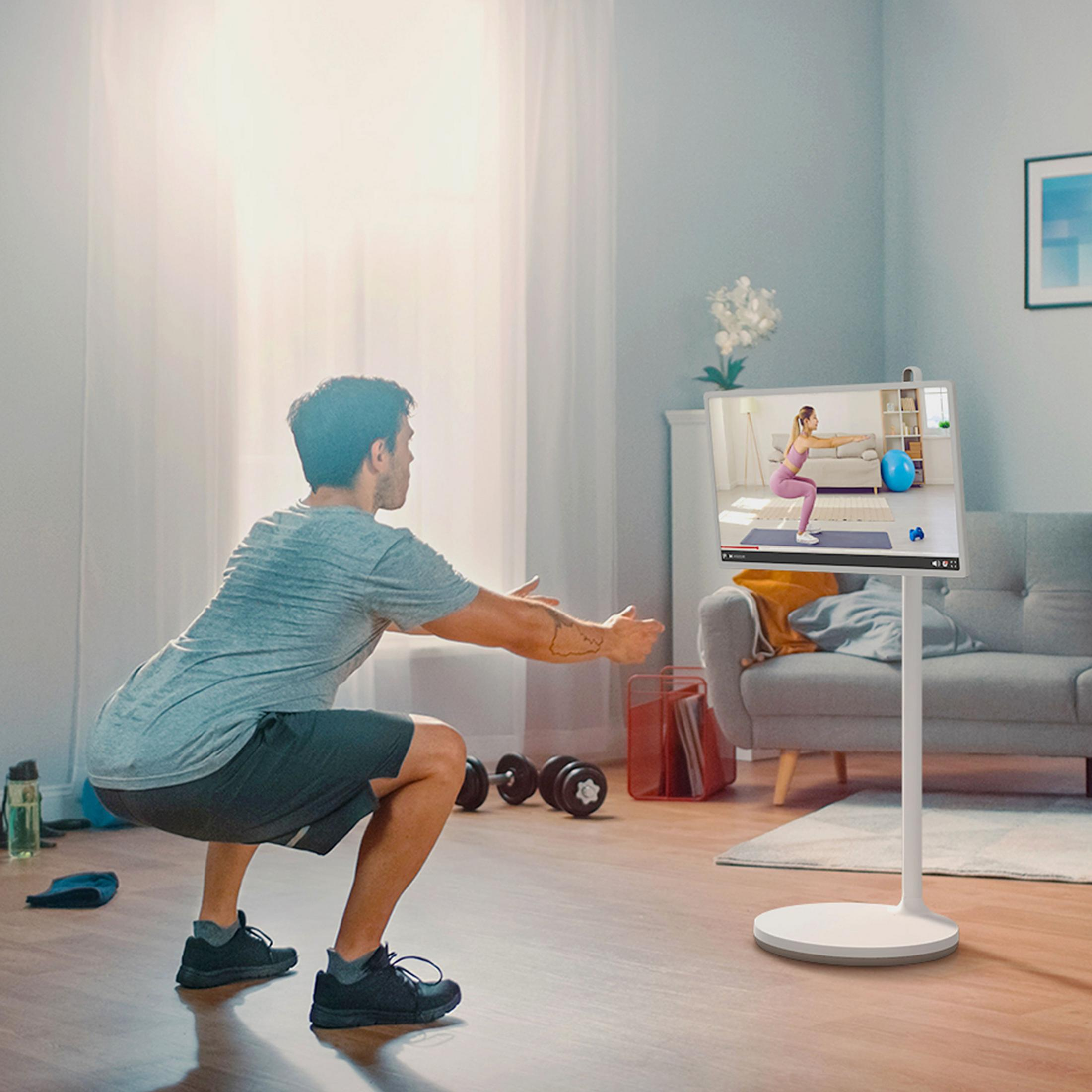 Un uomo fa squat a casa, seguendo un corso di fitness online su uno schermo.