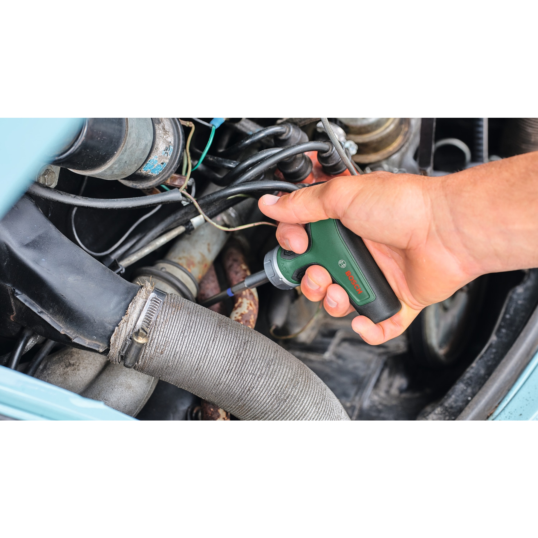 Eine Hand hält einen grünen Bosch Schrauber neben einem Auto Motor.