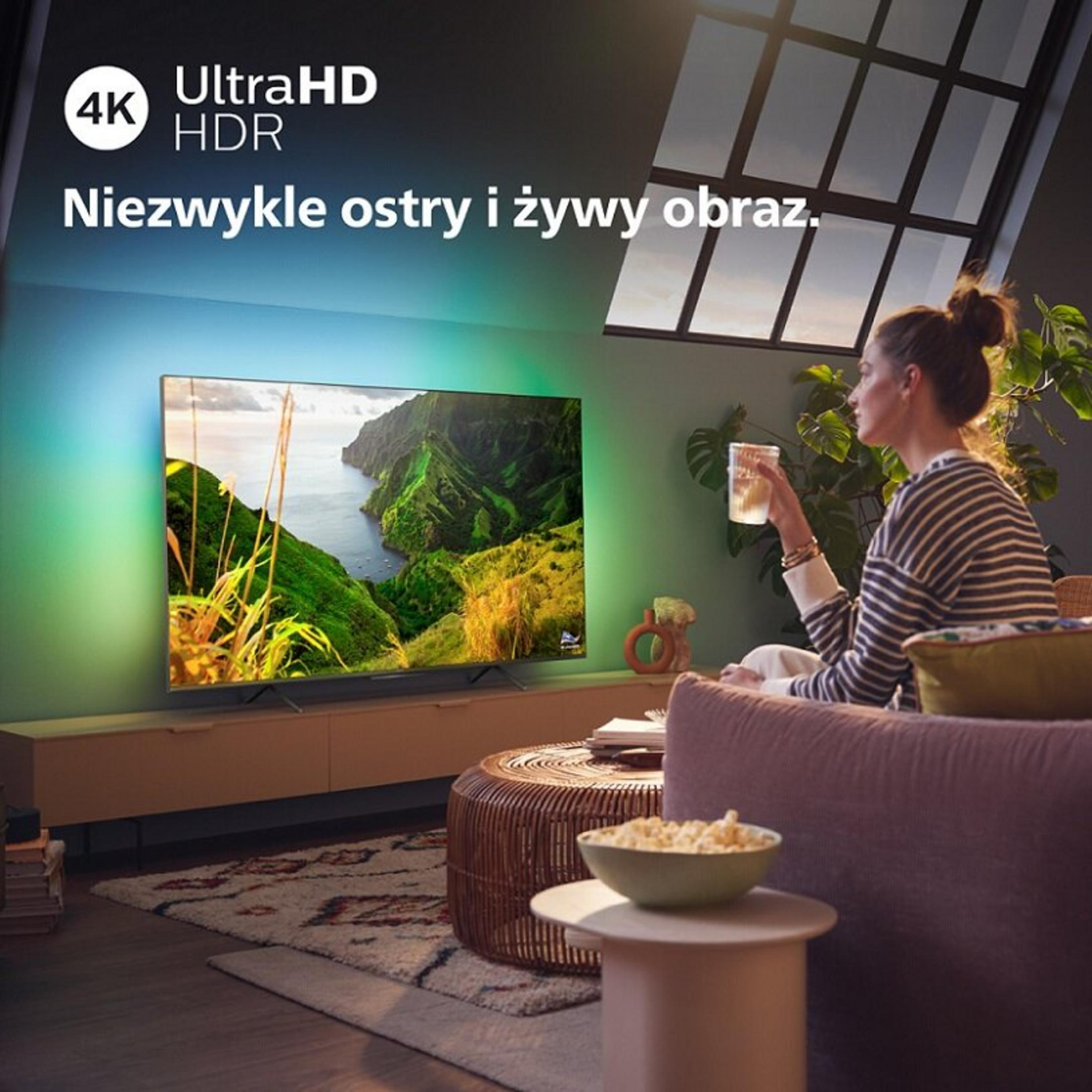 Mujer con jersey a rayas ve TV 4K con paisaje. Palomitas de maíz al lado.
