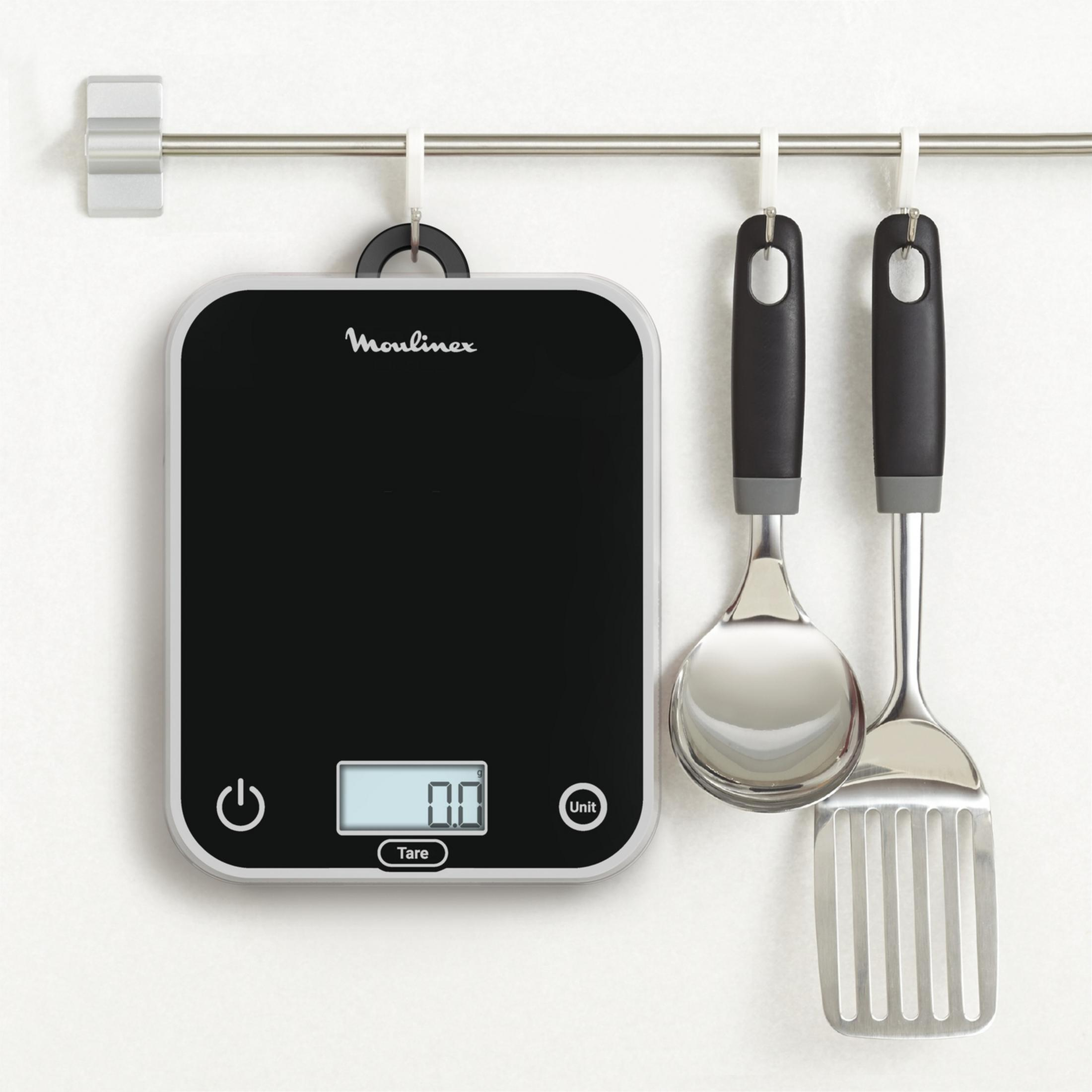 A black digital kitchen scale with two cooking utensils hangs on a wall rack.