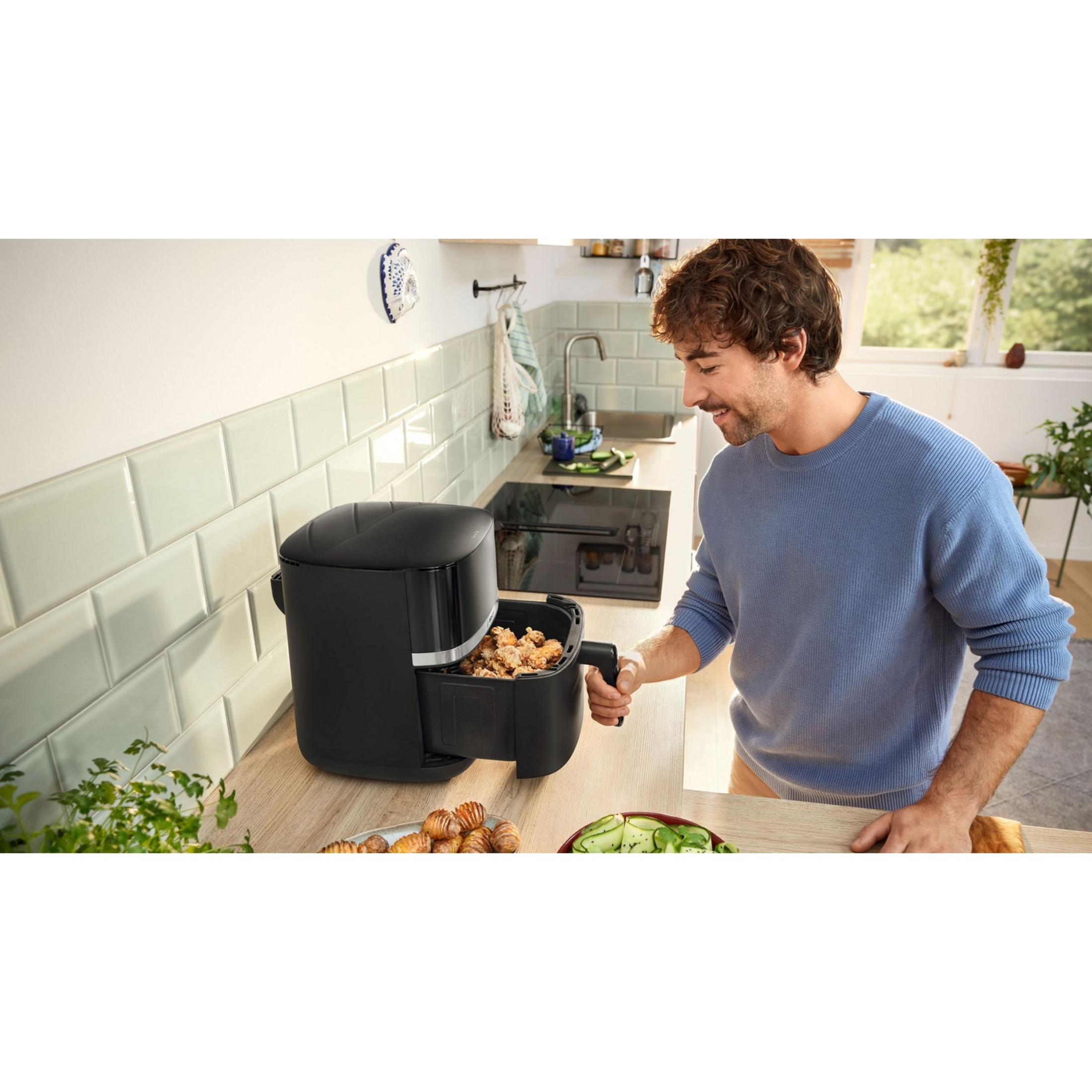 Un uomo con maglione blu controlla una friggitrice ad aria nera con cibo fritto.
