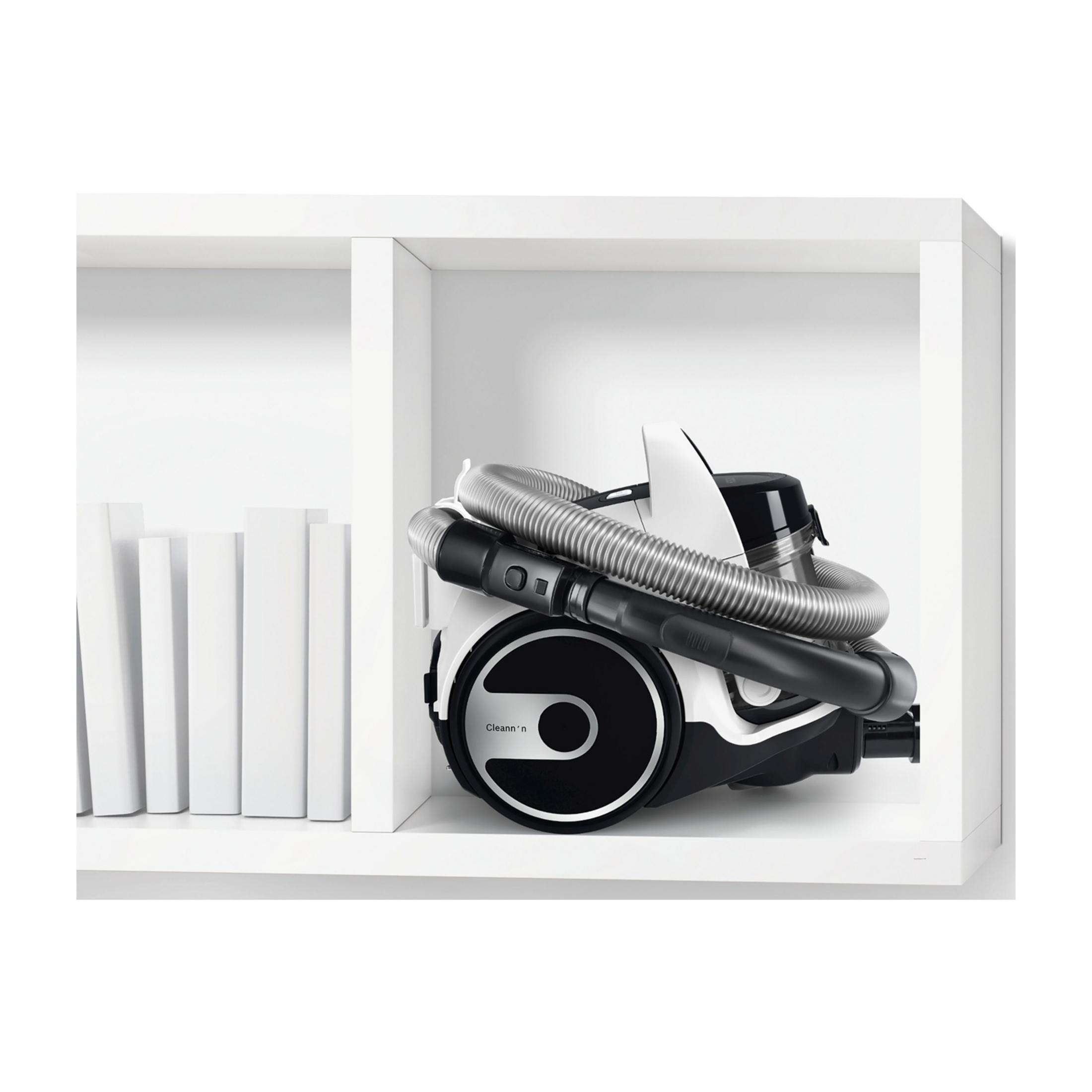 Biały odkurzacz z czarnym wężem leży na białej półce.