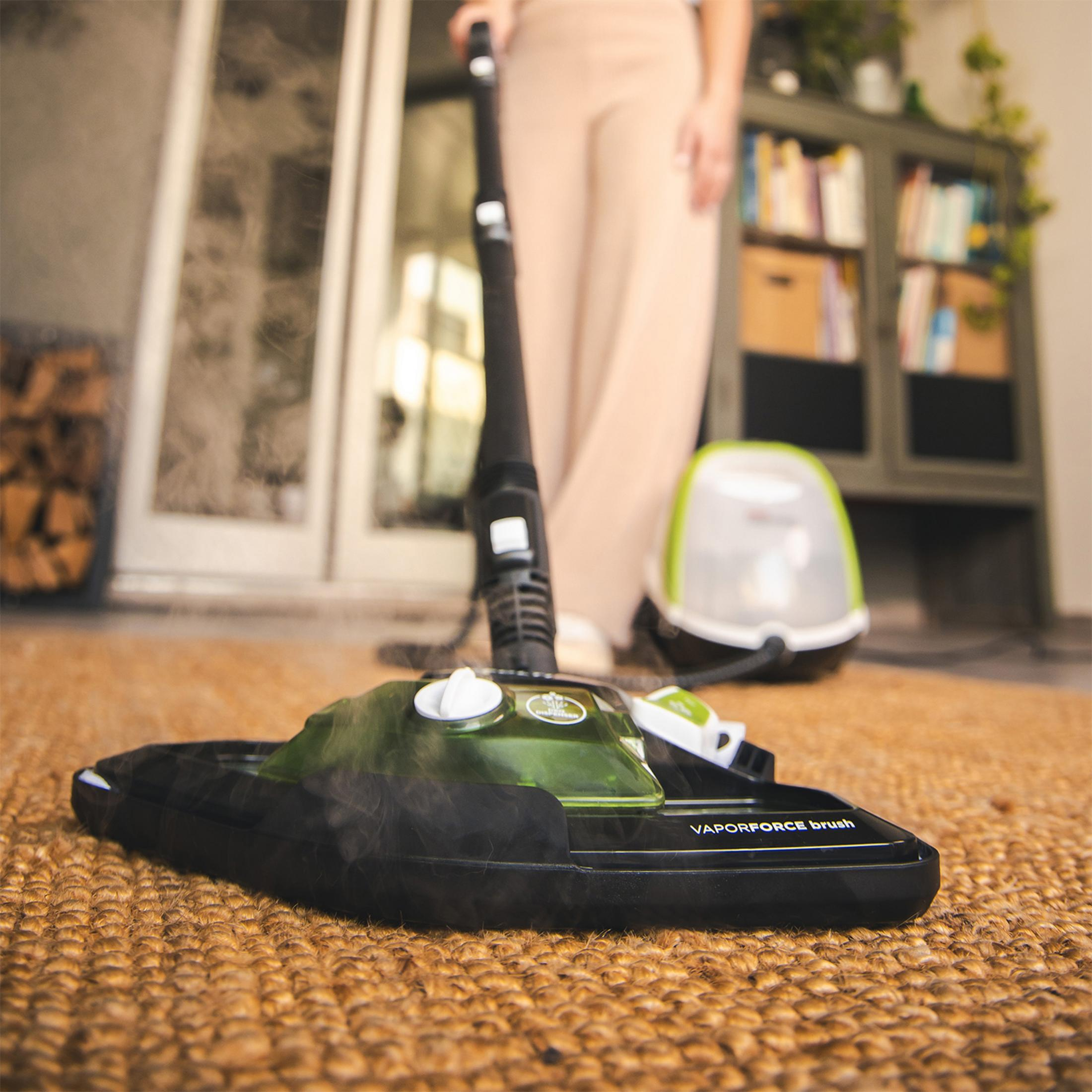 Szczotka parownicy na dywanie, unosząca się para. Osoba i urządzenie w tle.