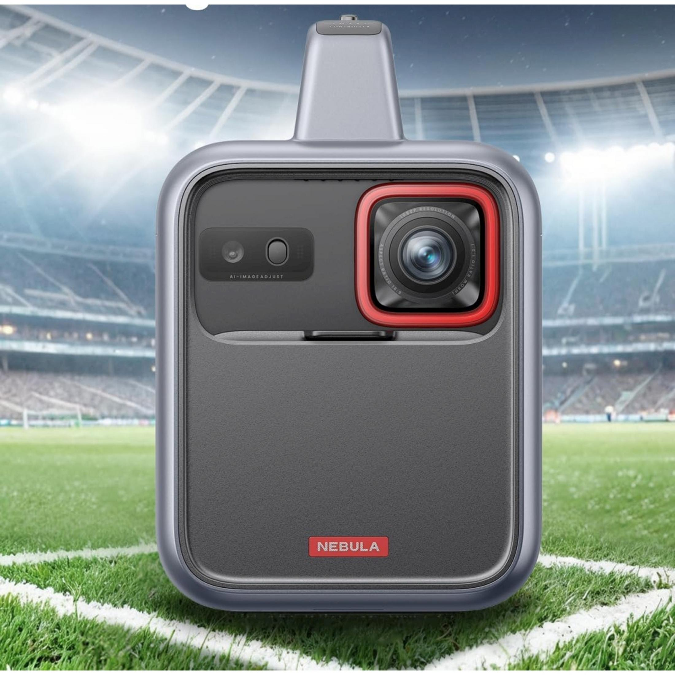 Szare urządzenie z obiektywem aparatu na boisku piłkarskim przed trybunami stadionu.