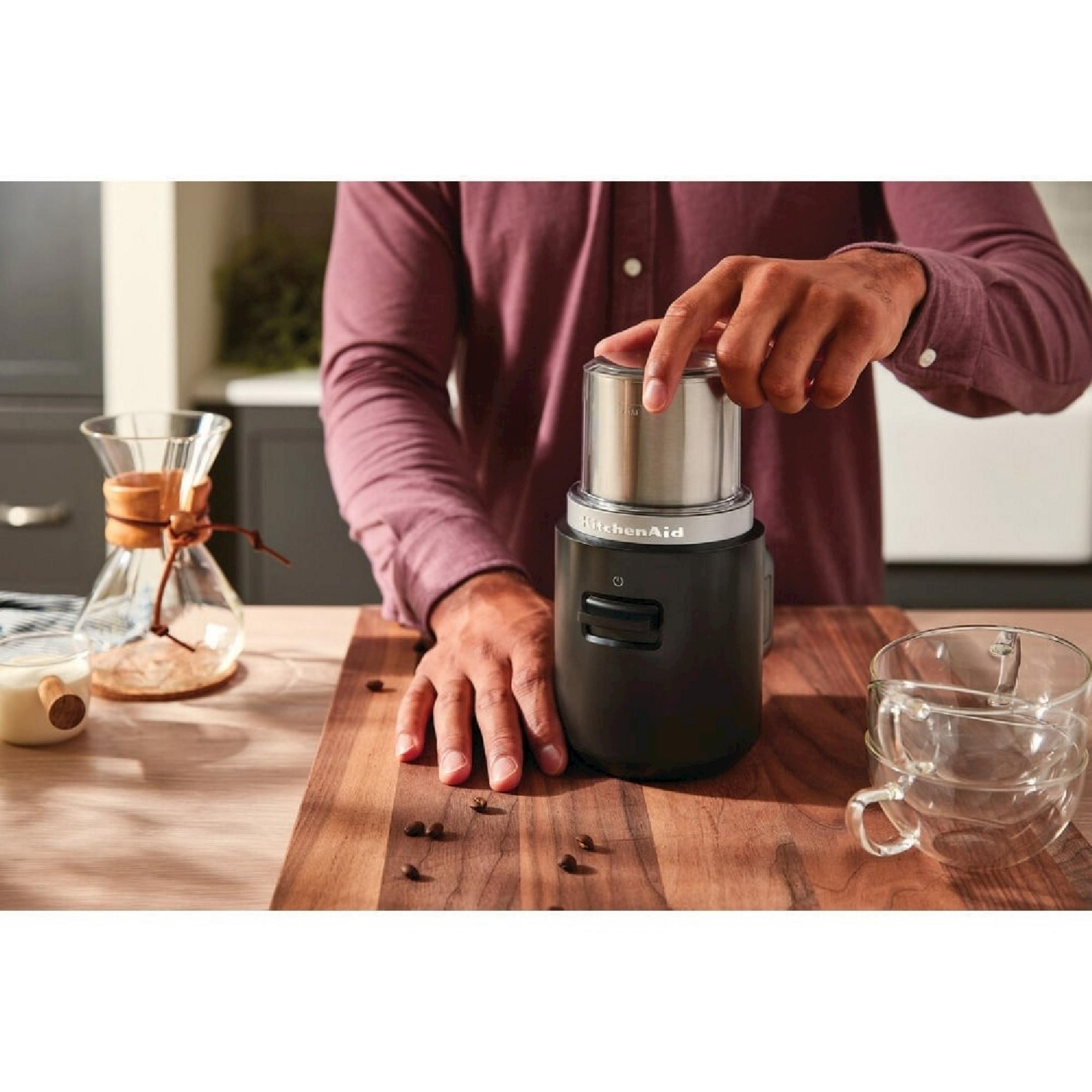Mann mahlt Kaffee mit KitchenAid-Mühle neben Chemex und Tassen auf einem Holzbrett.