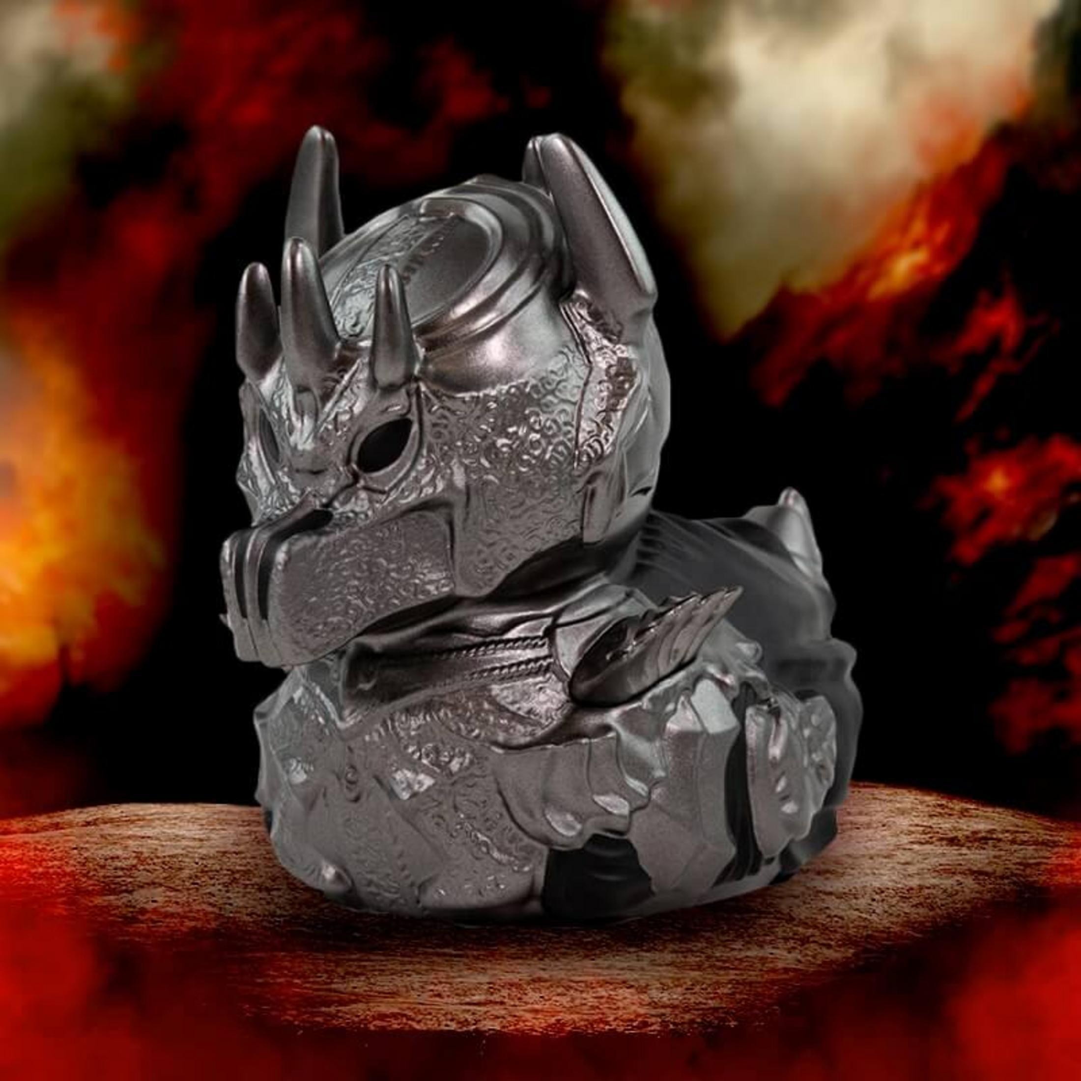 Eine silberne Gummiente in Drachenrüstung sitzt auf felsigem Boden vor einem feurigen Hintergrund.