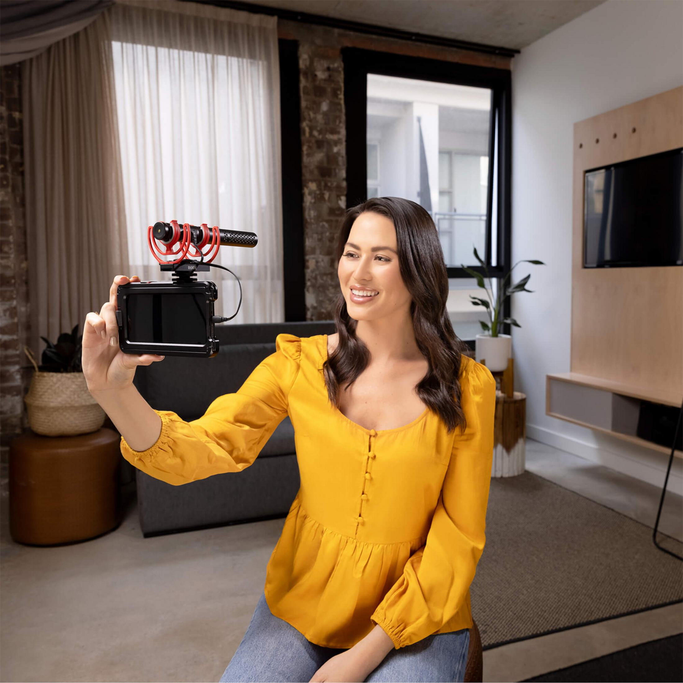 Kobieta w żółtej bluzce trzyma aparat z mikrofonem, filmuje się w salonie.