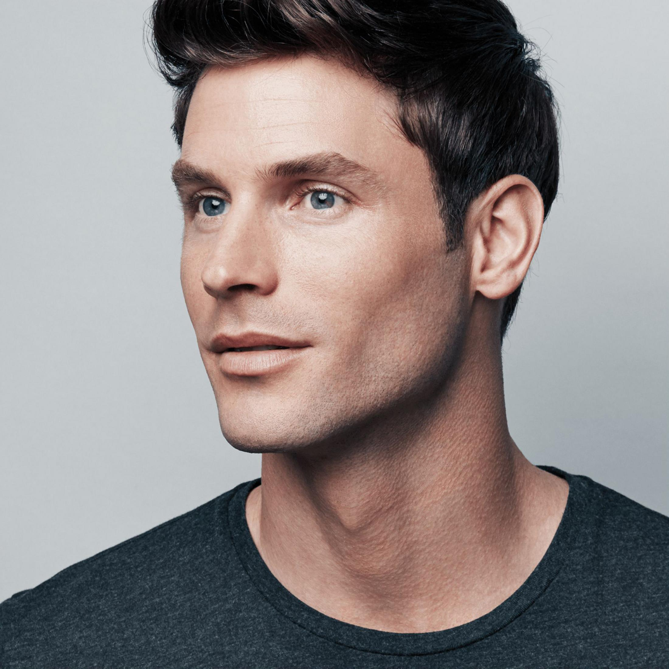 Mężczyzna z ciemnymi włosami i niebieskimi oczami nosi ciemnoszarą koszulkę.