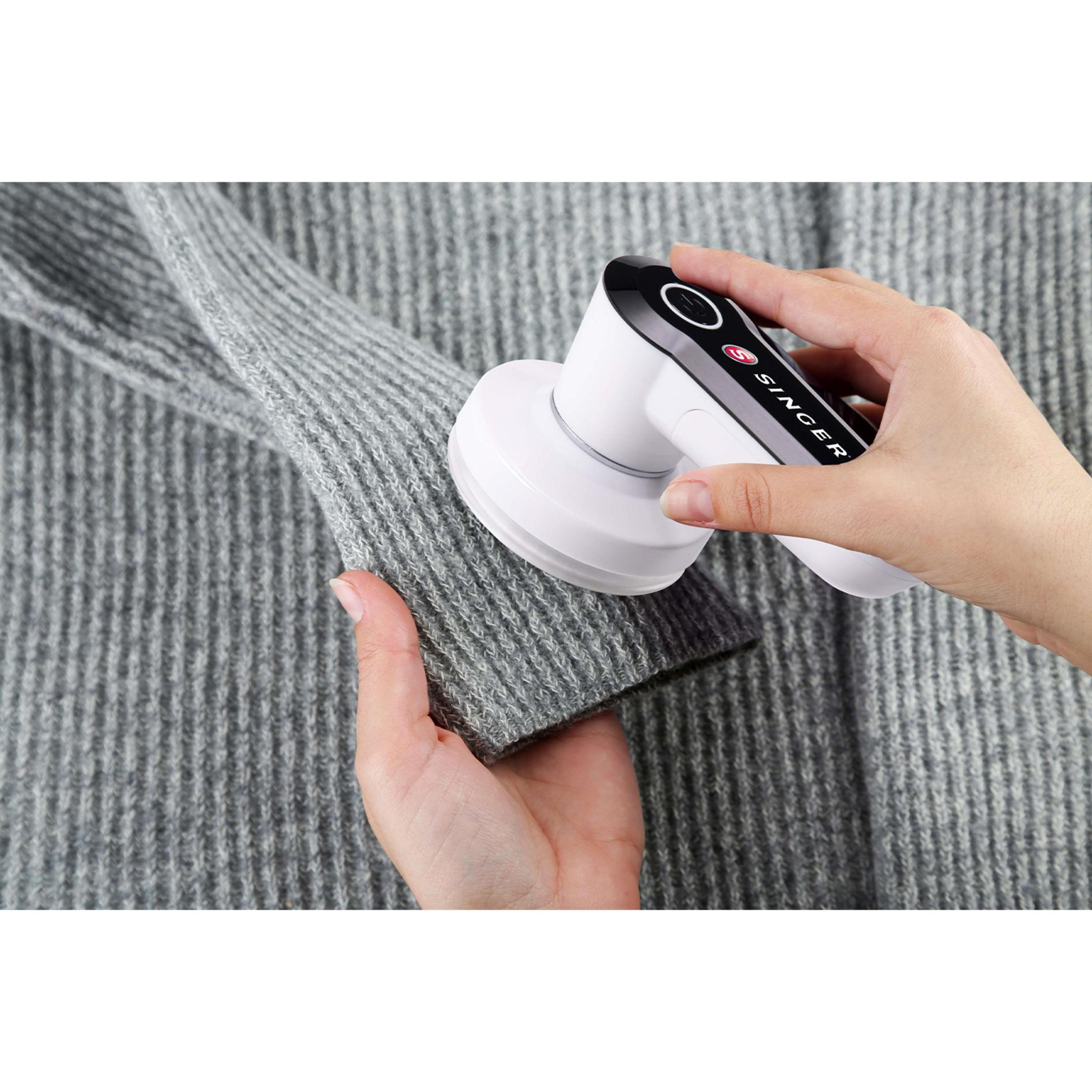 Eine Hand hält einen Fusselentferner über einen grauen Strickpullover.