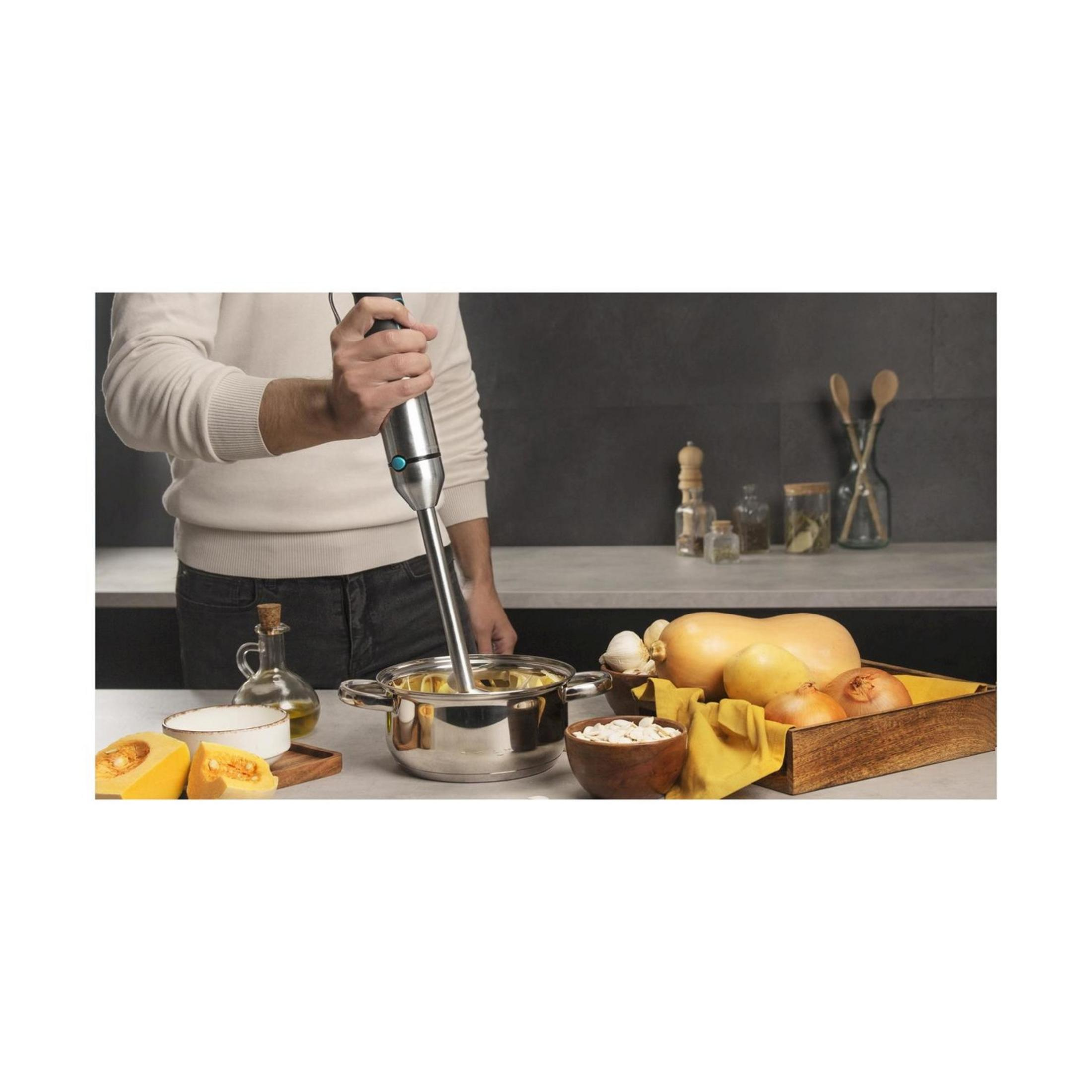 Een man mixt in een pan met een staafmixer. Ingrediënten zoals pompoen en uien liggen ernaast.
