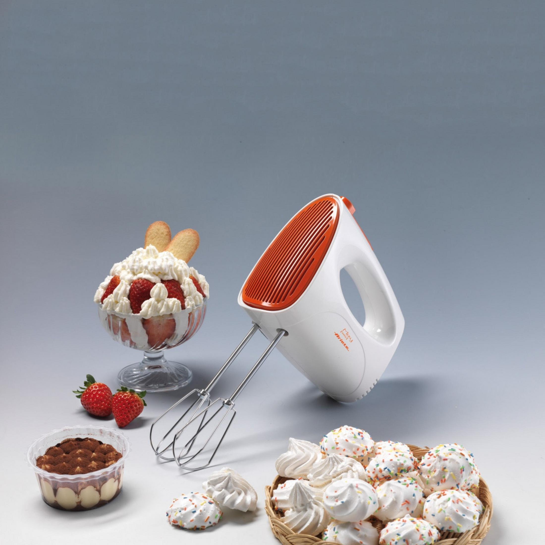Handmixer mit orangenem Oberteil neben Desserts: Tiramisu, Erdbeeren und Baiser.