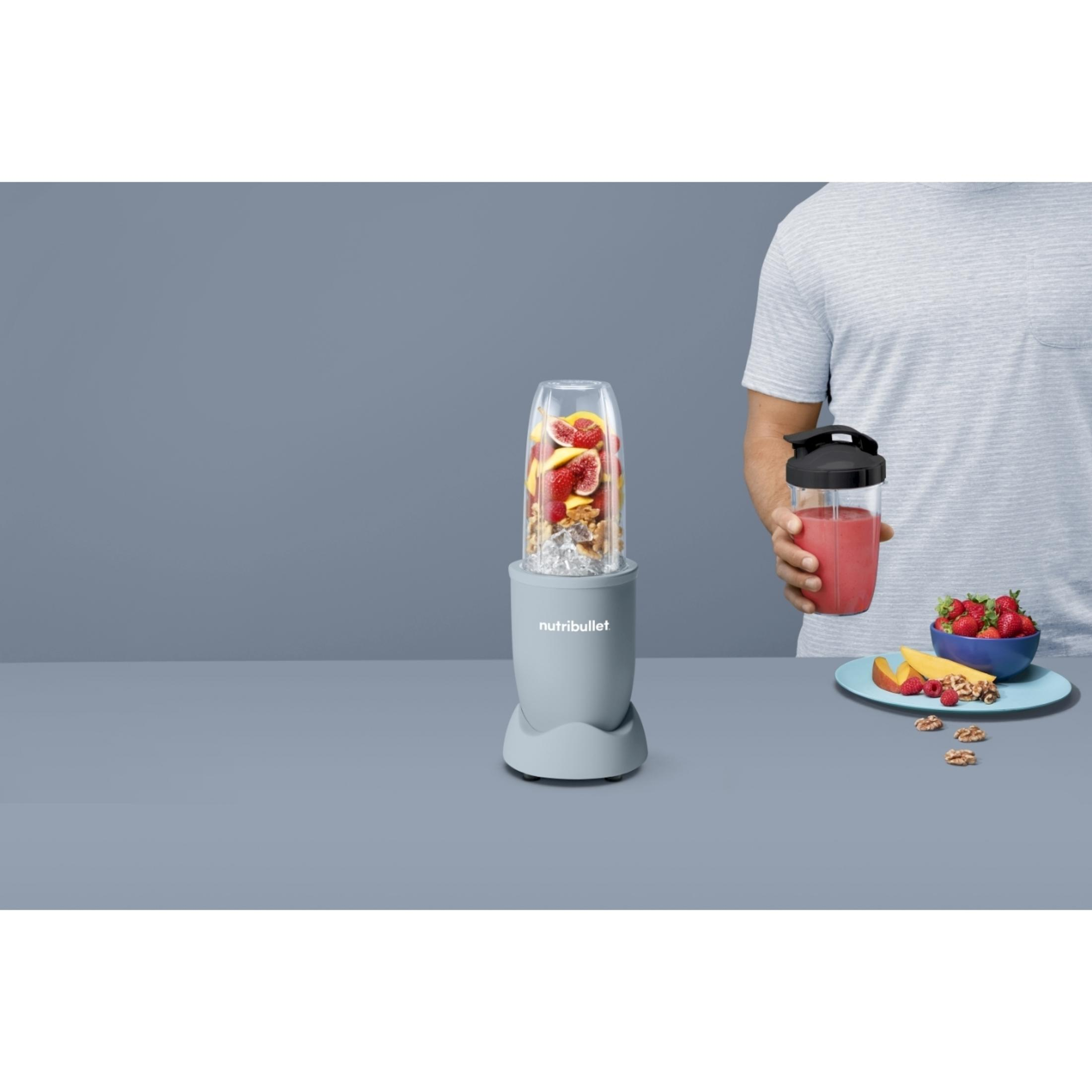 Un homme tient une tasse à smoothie. Un mixeur avec des ingrédients à côté.