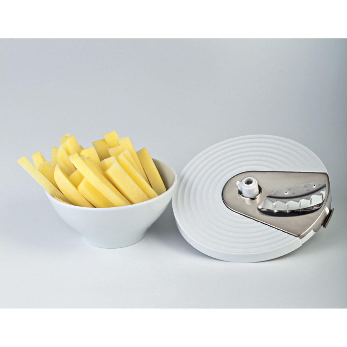 Un bol blanco con patatas cortadas se sitúa junto a una cortadora de patatas blanca y plateada.