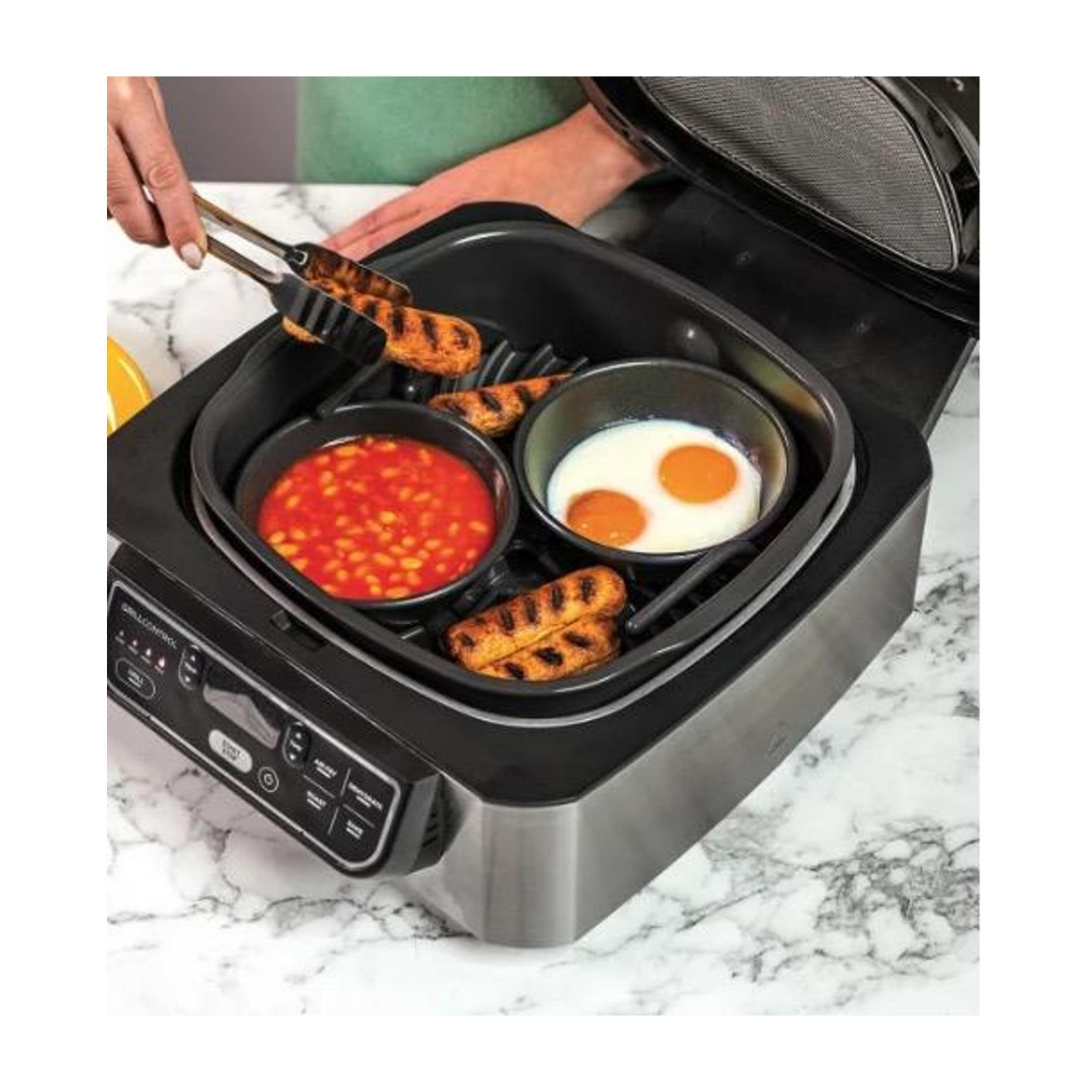 Würstchen werden mit Zange in einen Grill gegeben, daneben Bohnen und Eier.