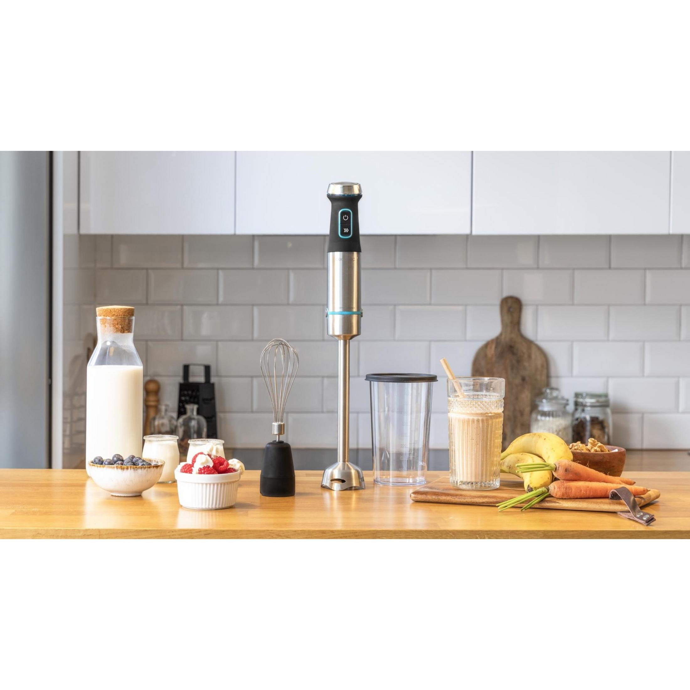 Una batidora de inmersión con accesorios, fruta y leche en mesa de madera.