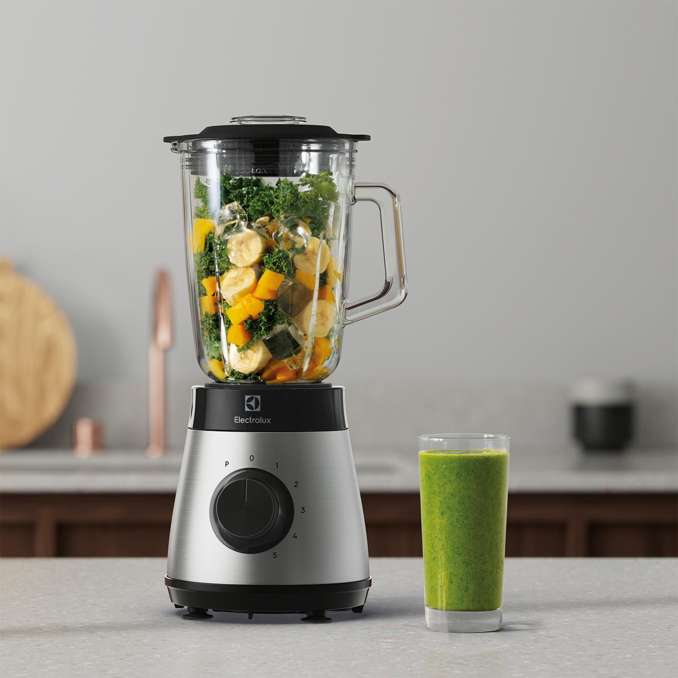 Una licuadora plateada con frutas y verduras, al lado un vaso de batido verde.