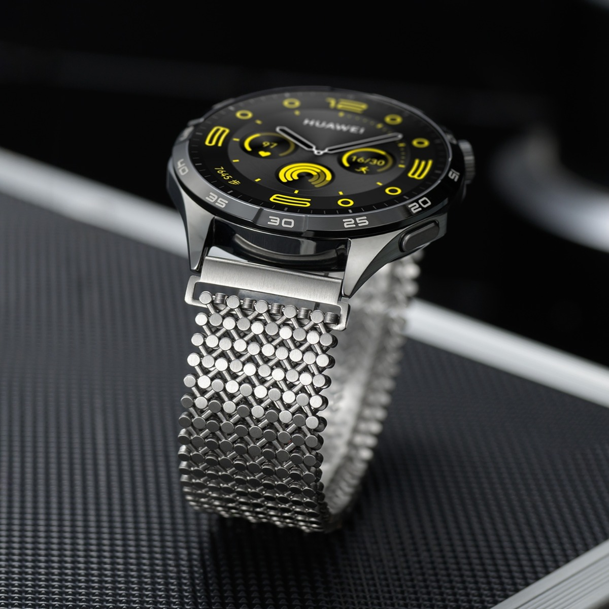 Uno smartwatch con quadrante nero, accenti gialli e cinturino in maglia argentata.