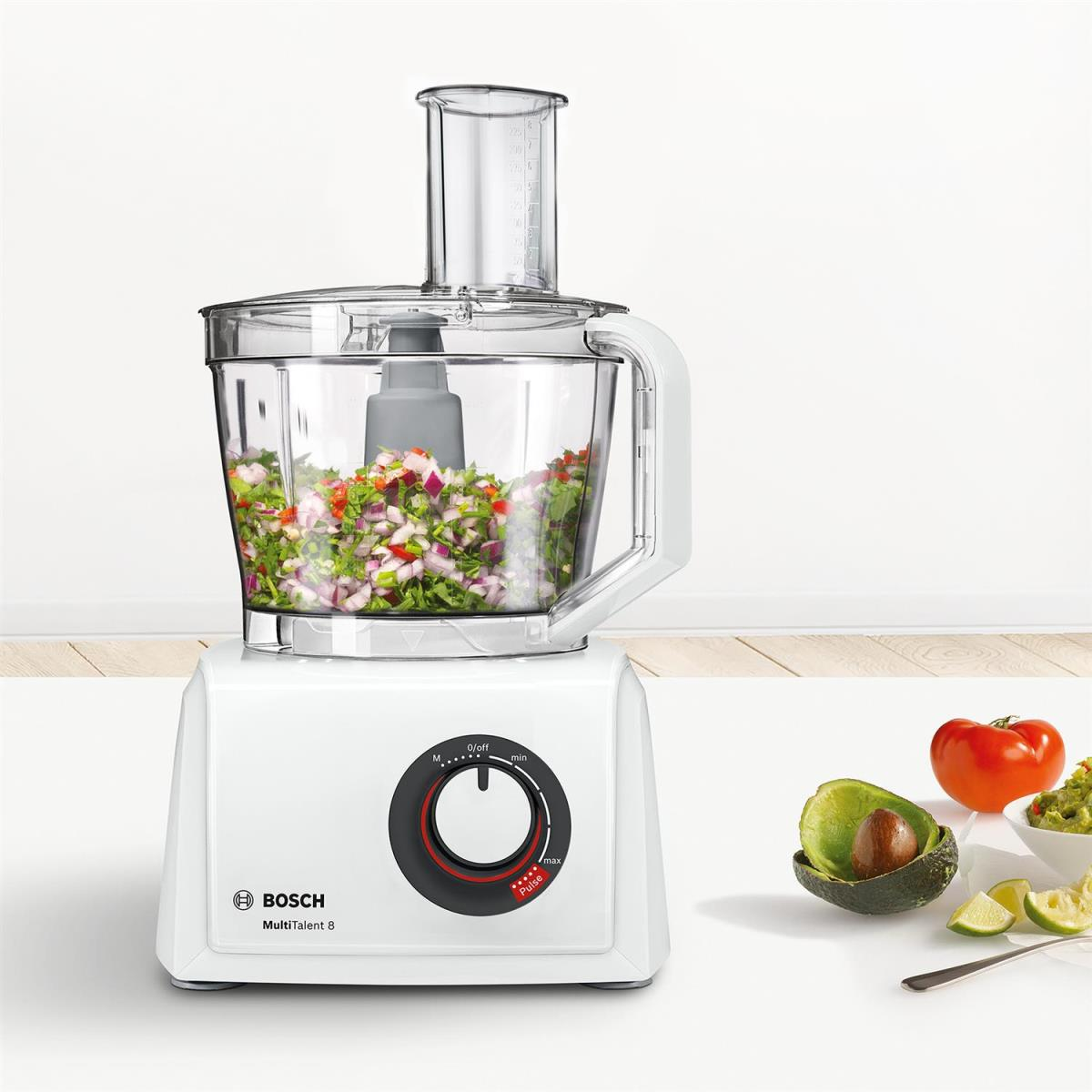 Ein weißer Bosch Küchenmaschinen-Aufsatz mit gehacktem Gemüse und daneben Avocado und Tomate.