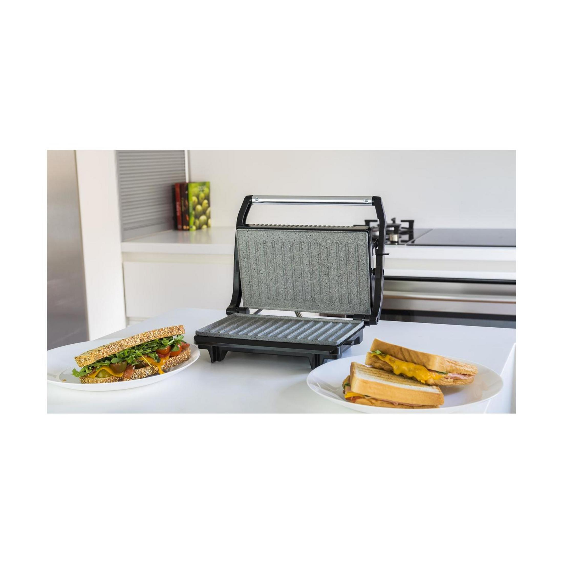 Otwarty opiekacz do kanapek stoi na blacie z dwoma talerzami kanapek obok.