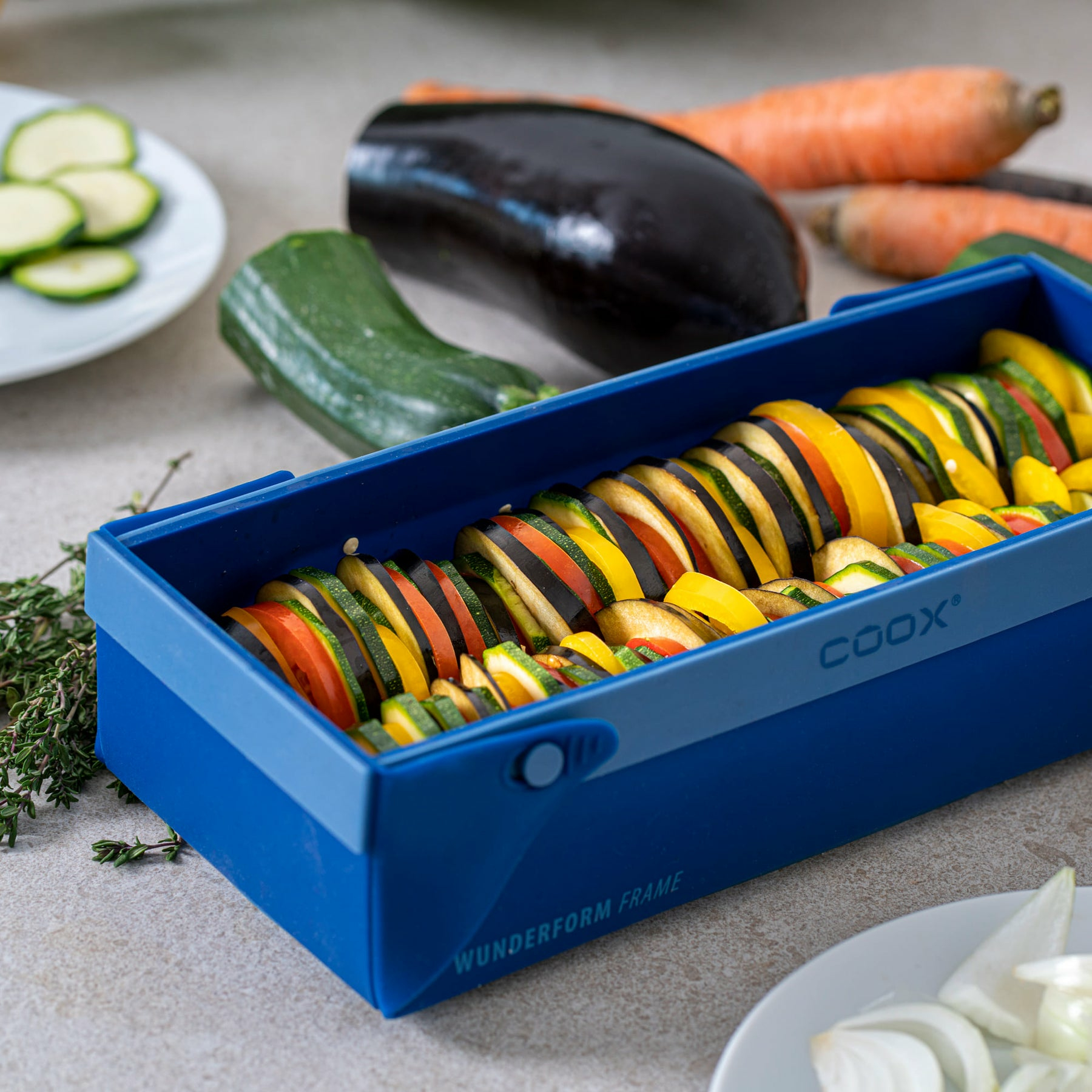 Gemüsescheiben in einem blauen Behälter, daneben Zucchini, Aubergine und Karotten.