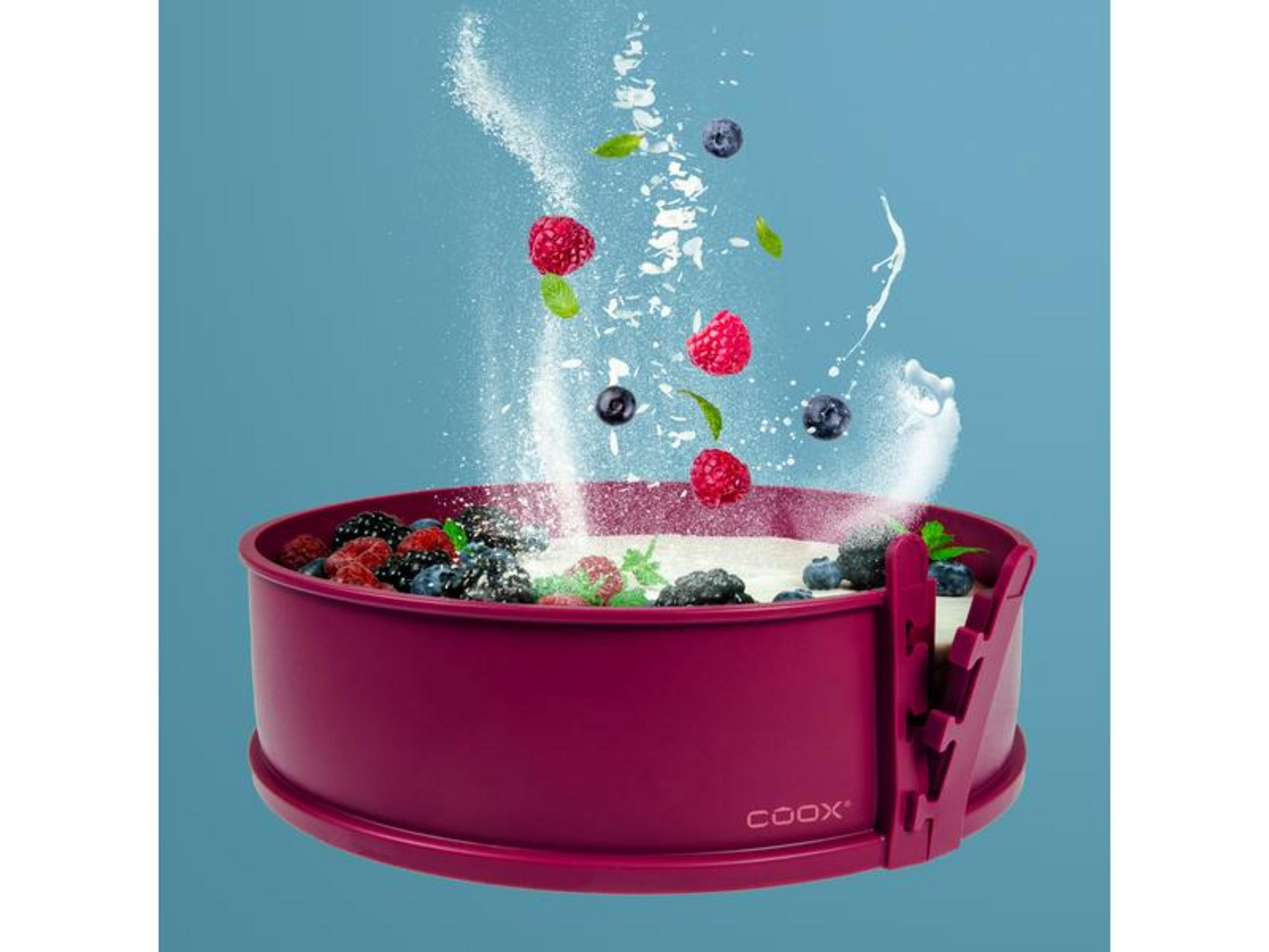 Himbeeren, Blaubeeren und Staubzucker fallen in eine bordeauxrote Kuchenform.