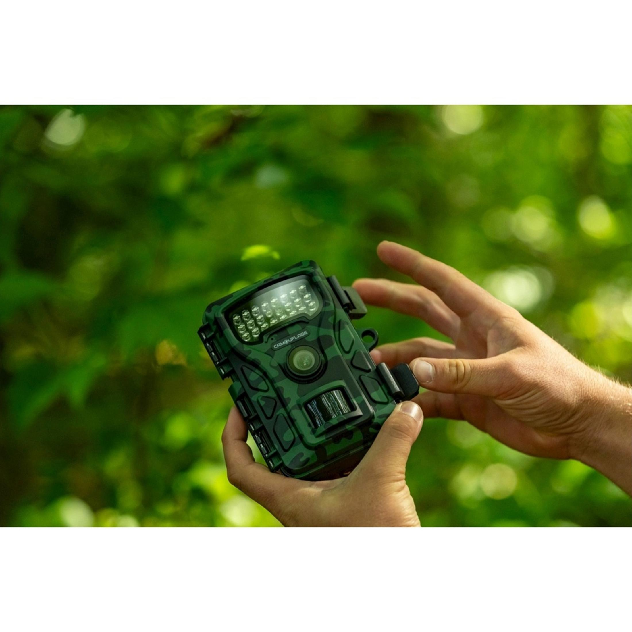 Fototrappola mimetica verde con molti LED e lente, tenuta in mano, sfondo sfocato.