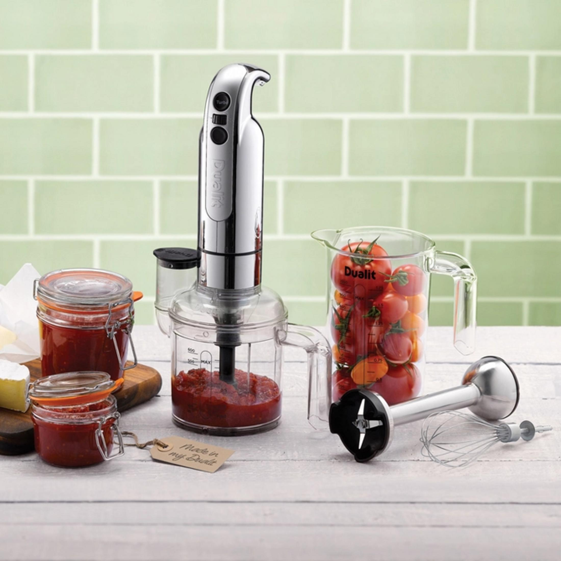 Ein Stabmixer mit Zubehör, Marmelade und Tomaten vor einer grünen Kachelwand.