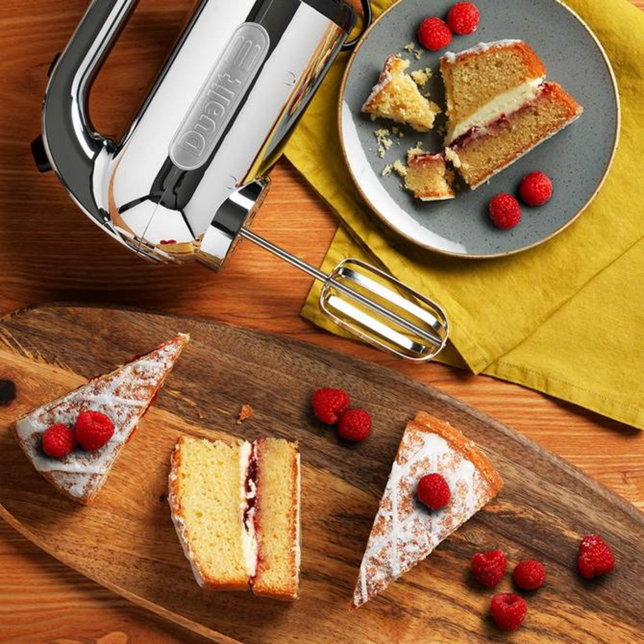 Un batteur à main argenté à côté de parts de gâteau et de framboises.