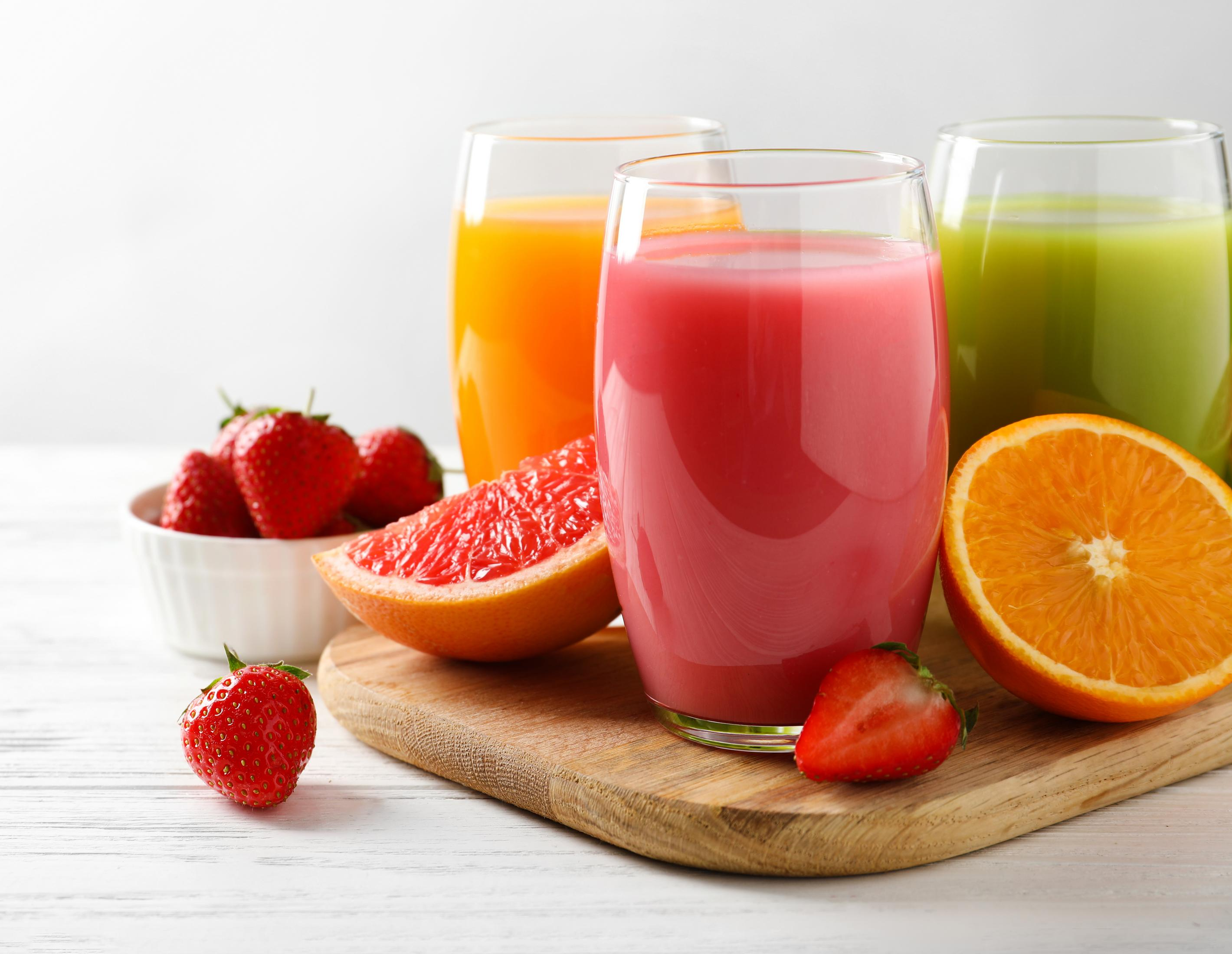 Drei Gläser Saft (orange, rosa, grün) mit Früchten auf Holzbrett.