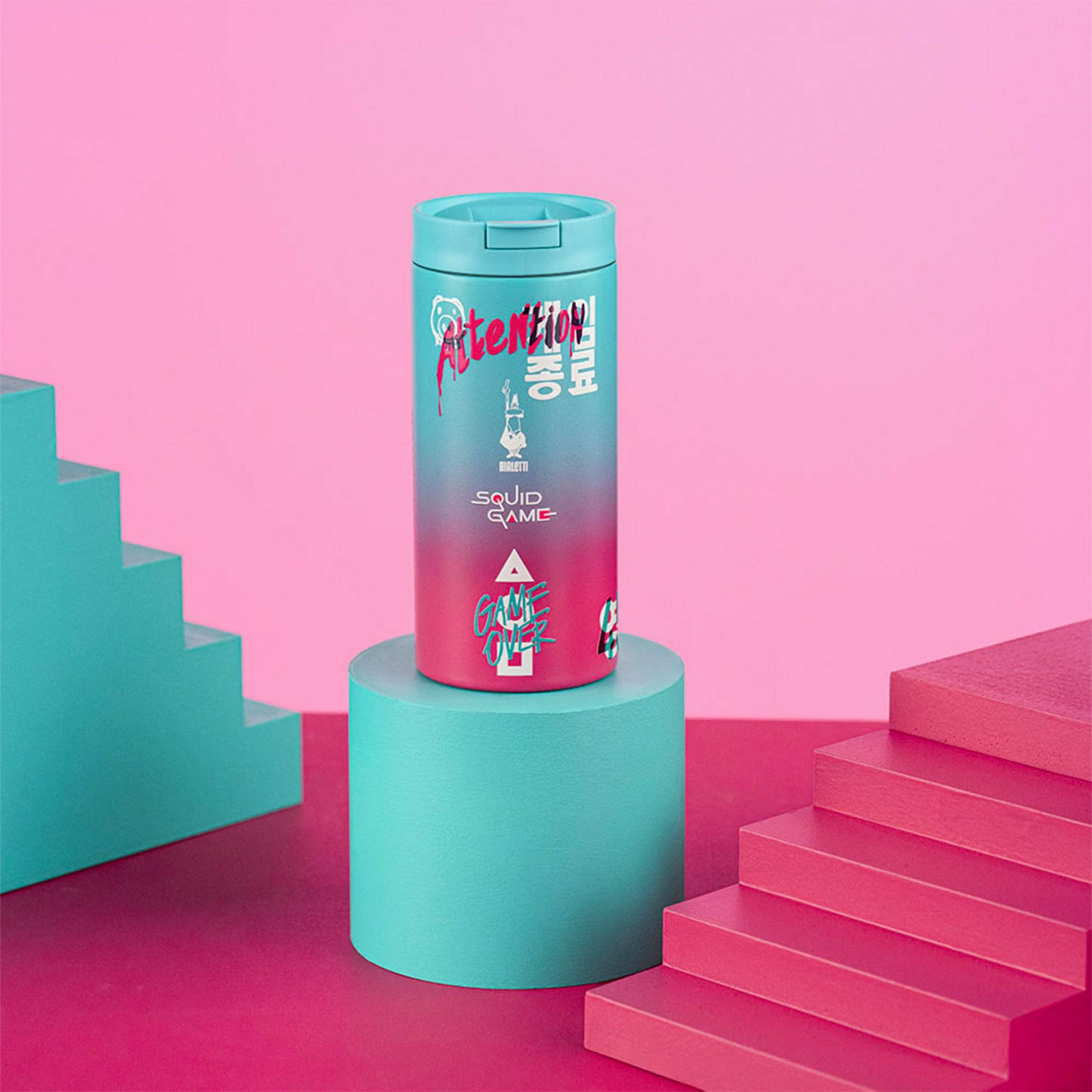 Una tazza blu con testo "Squid Game" si trova su un piedistallo turchese.