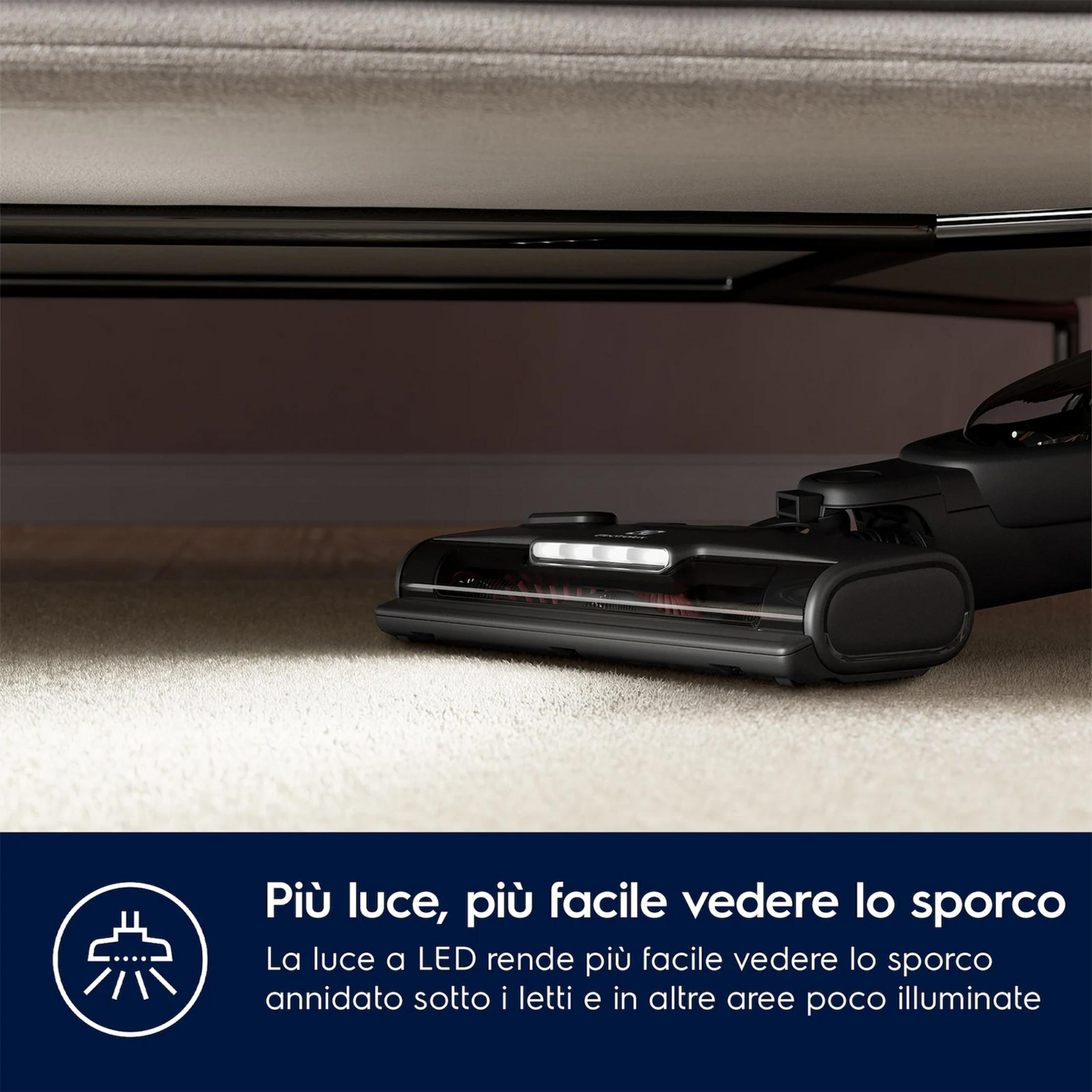 Cepillo aspirador con luz LED debajo de una cama sobre alfombra clara.
