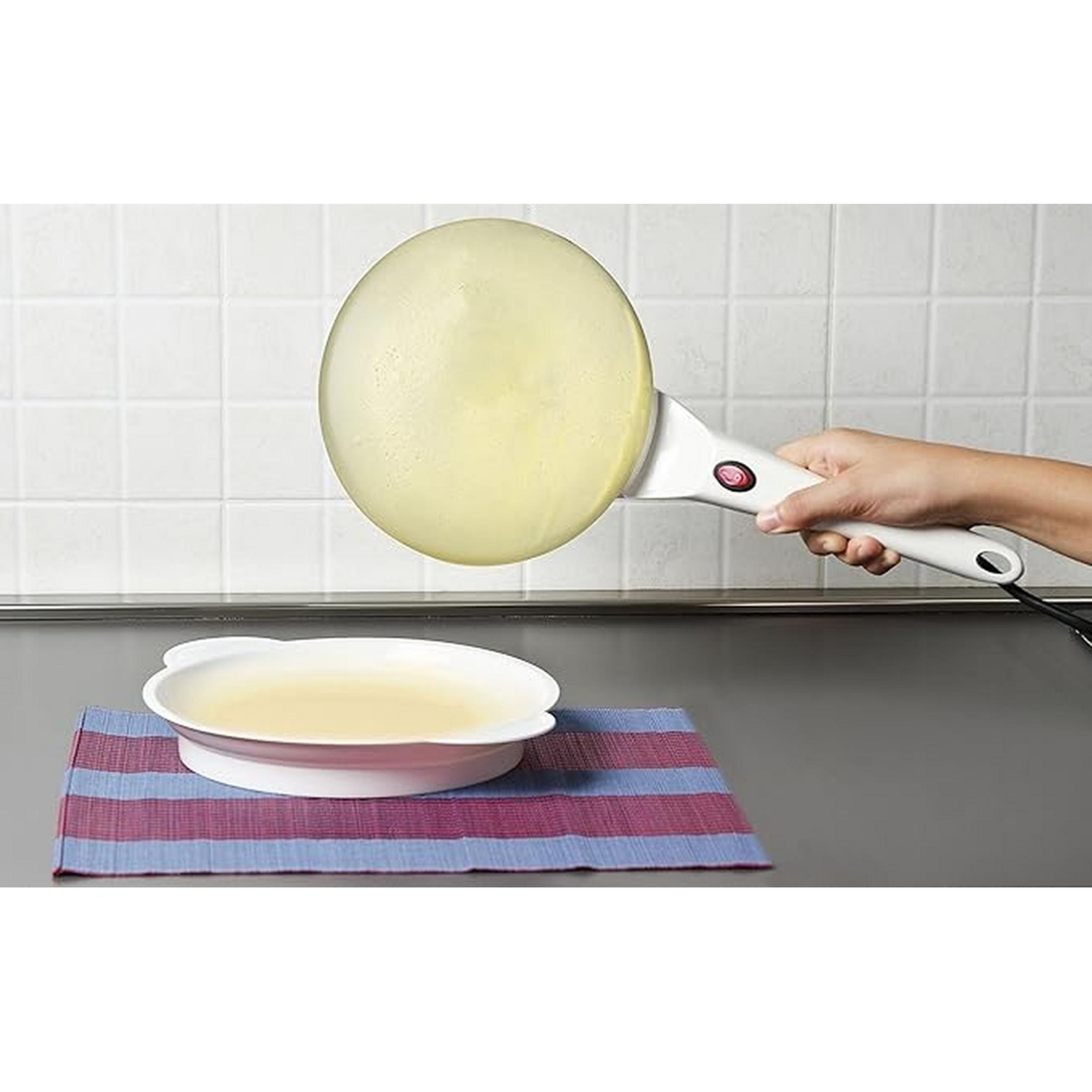 Een hand houdt een witte crêpemaker vast met geel beslag. Een kom ernaast.