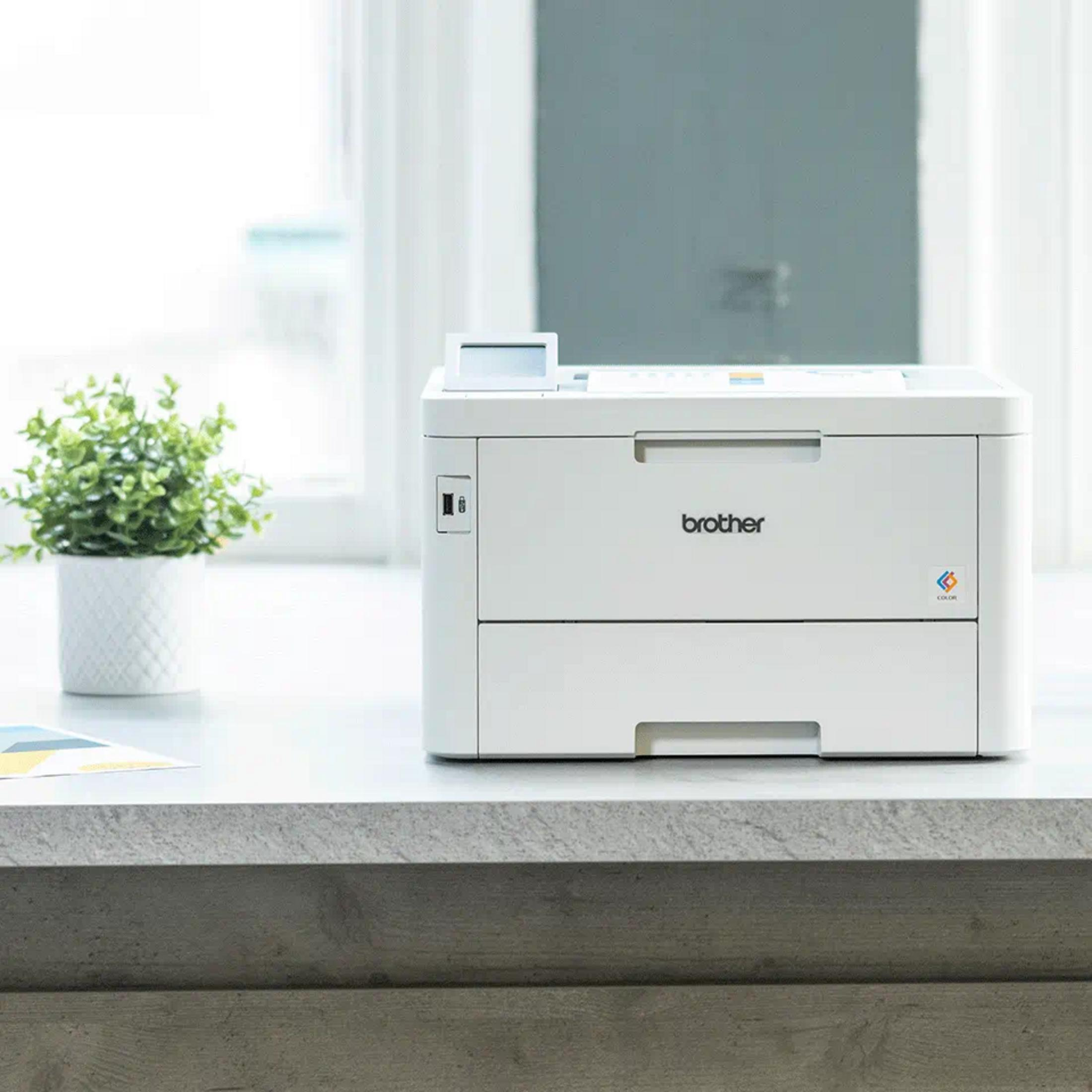 Une imprimante couleur Brother blanche est sur un bureau gris à côté d'une plante en pot.