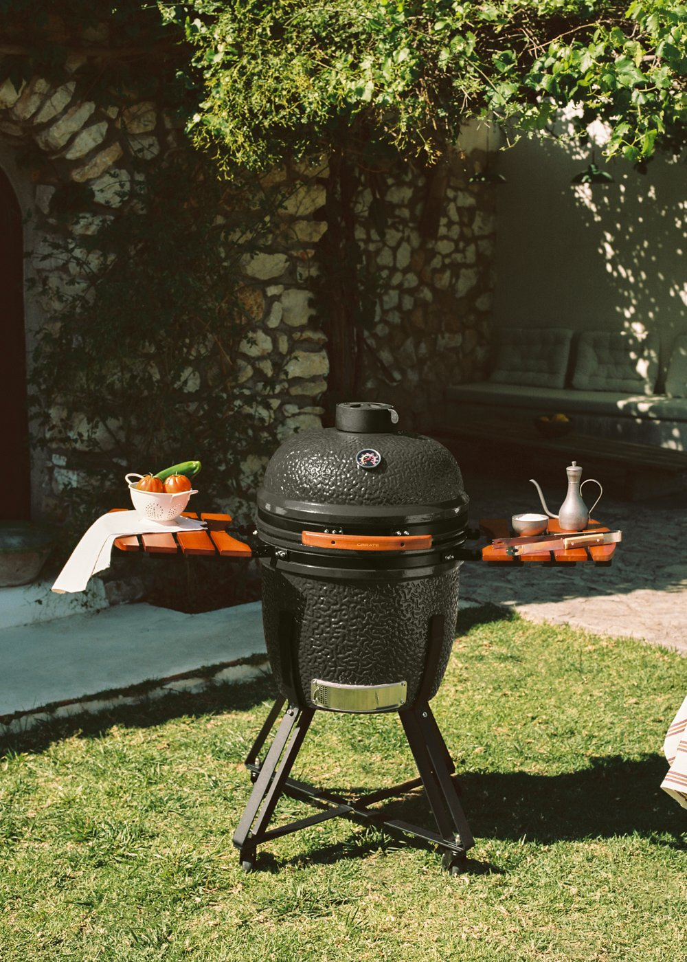 Parrilla negra de carbón en césped con tomates y pepino en mesa.