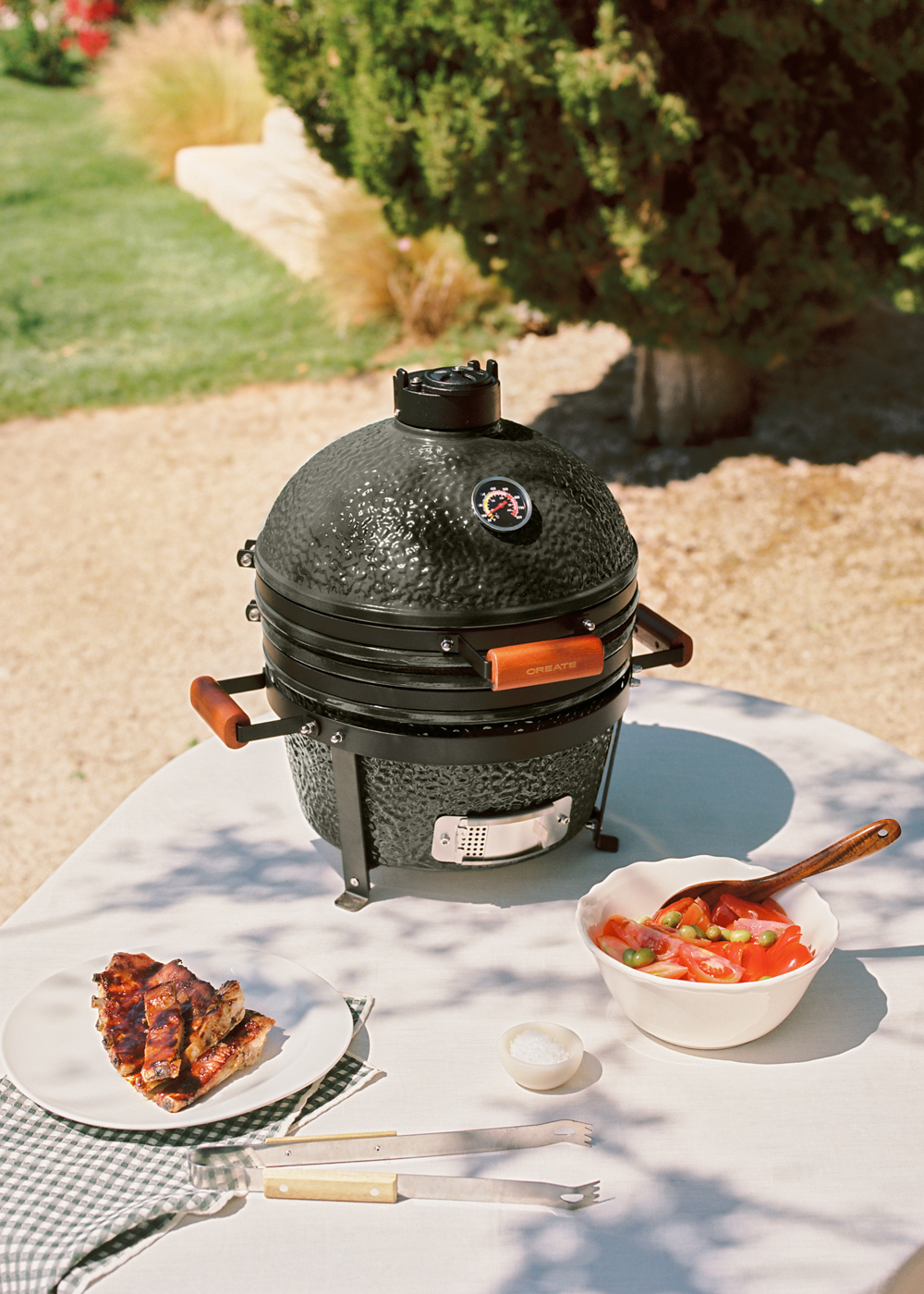 Una parrilla negra con asas de madera sobre una mesa exterior con carne a la parrilla y ensalada.