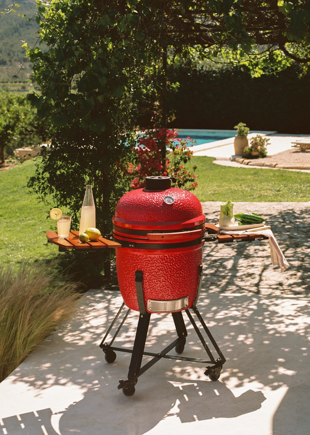 Una parrilla de cerámica roja se asienta en un patio sombreado con vistas a un jardín y piscina.
