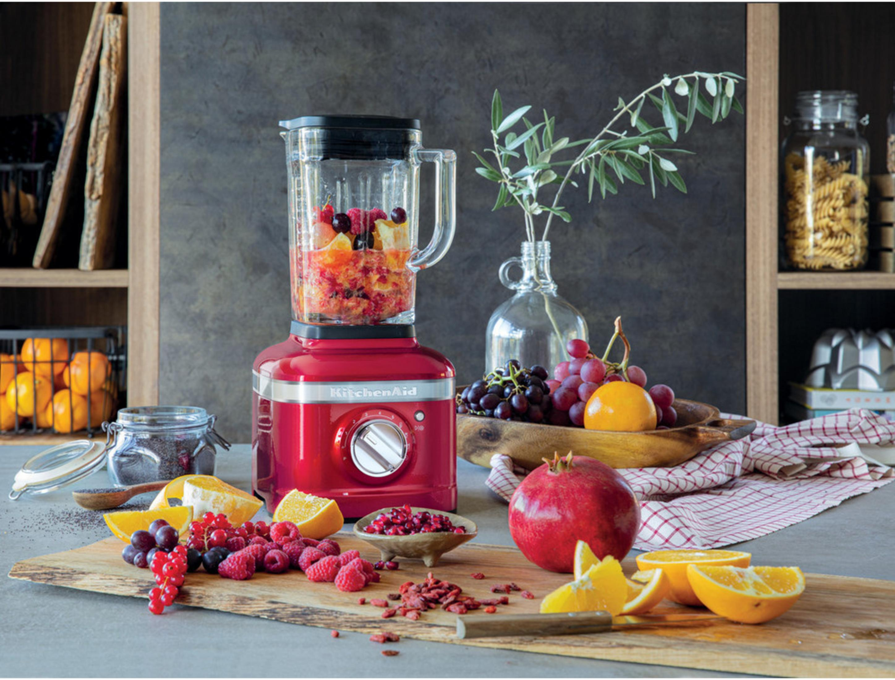 Ein roter Mixer mit Früchten, daneben Beeren, Orangen und Trauben.