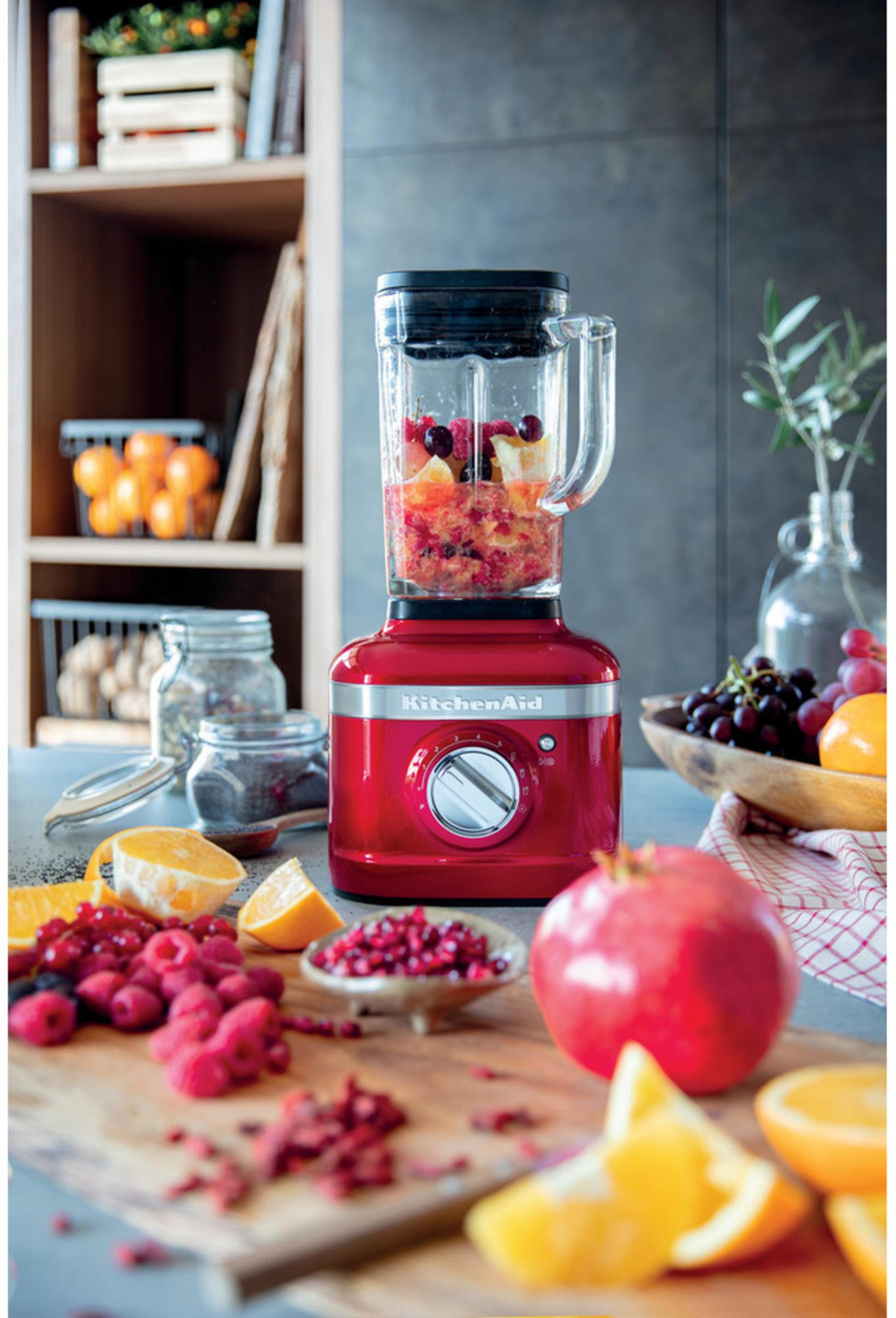Ein roter Standmixer mit Früchten gefüllt, daneben frische Beeren, Orangen und Granatapfel.