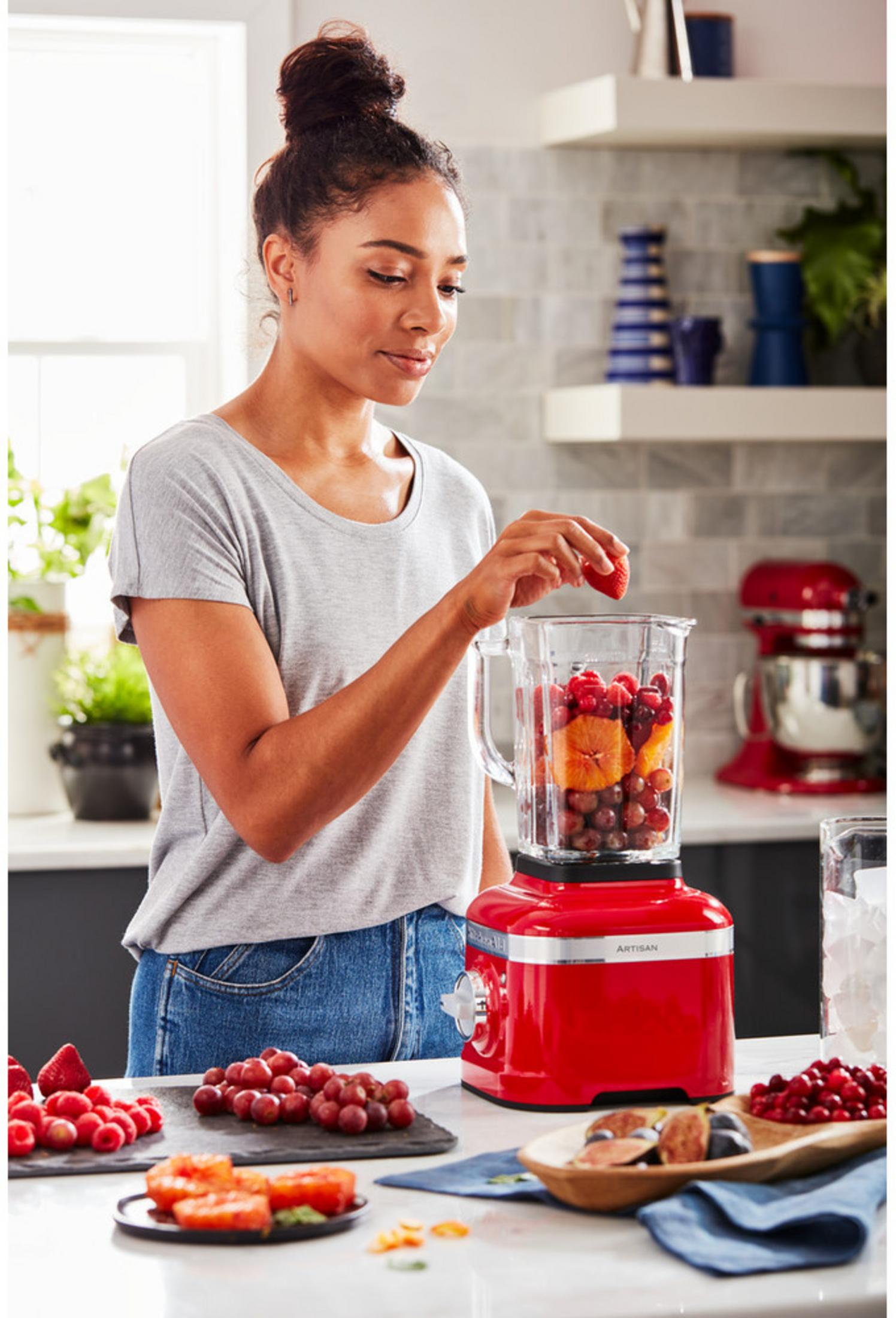 Frau gibt Erdbeere in roten Mixer mit Obst. Früchte liegen auf dem Tisch.