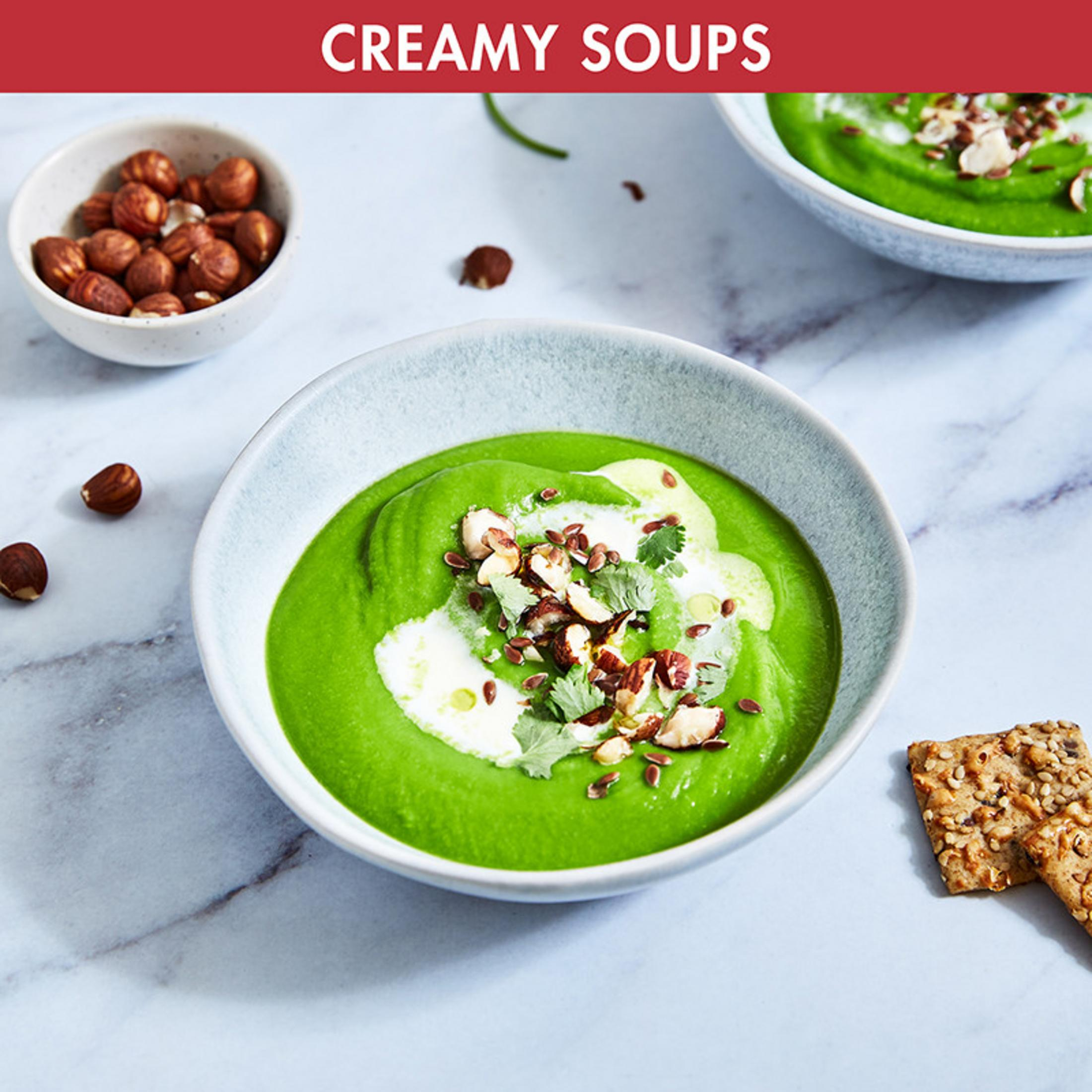 Grüne Suppe garniert mit Nüssen und Samen in einer Schüssel, Haselnüsse und Kekse daneben.