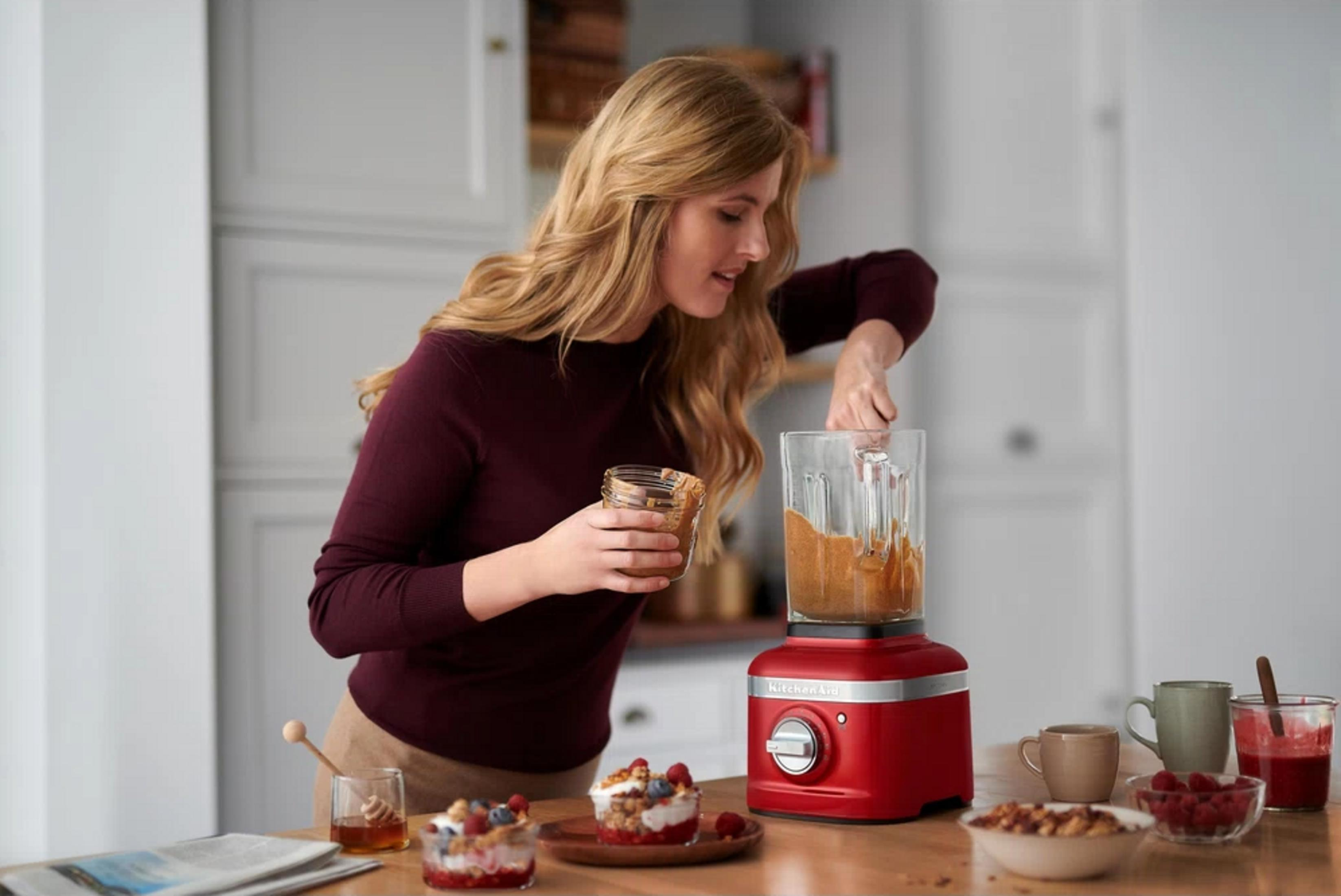 Eine Frau gibt Mandelbutter in einen roten Mixer. Frühstück und Beeren liegen daneben.