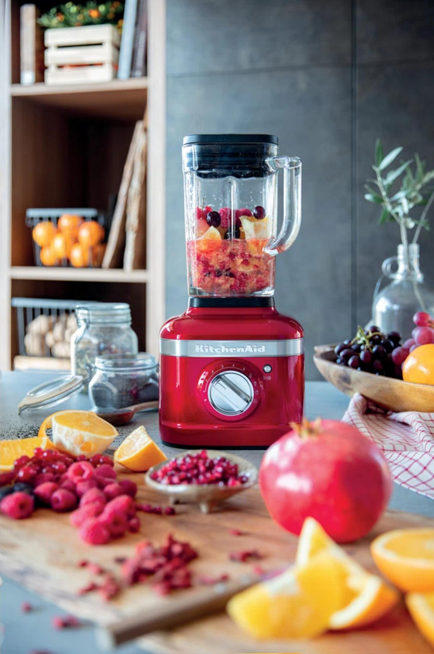Ein roter Standmixer mit Früchten, daneben Beeren, Orangen und Granatapfel.