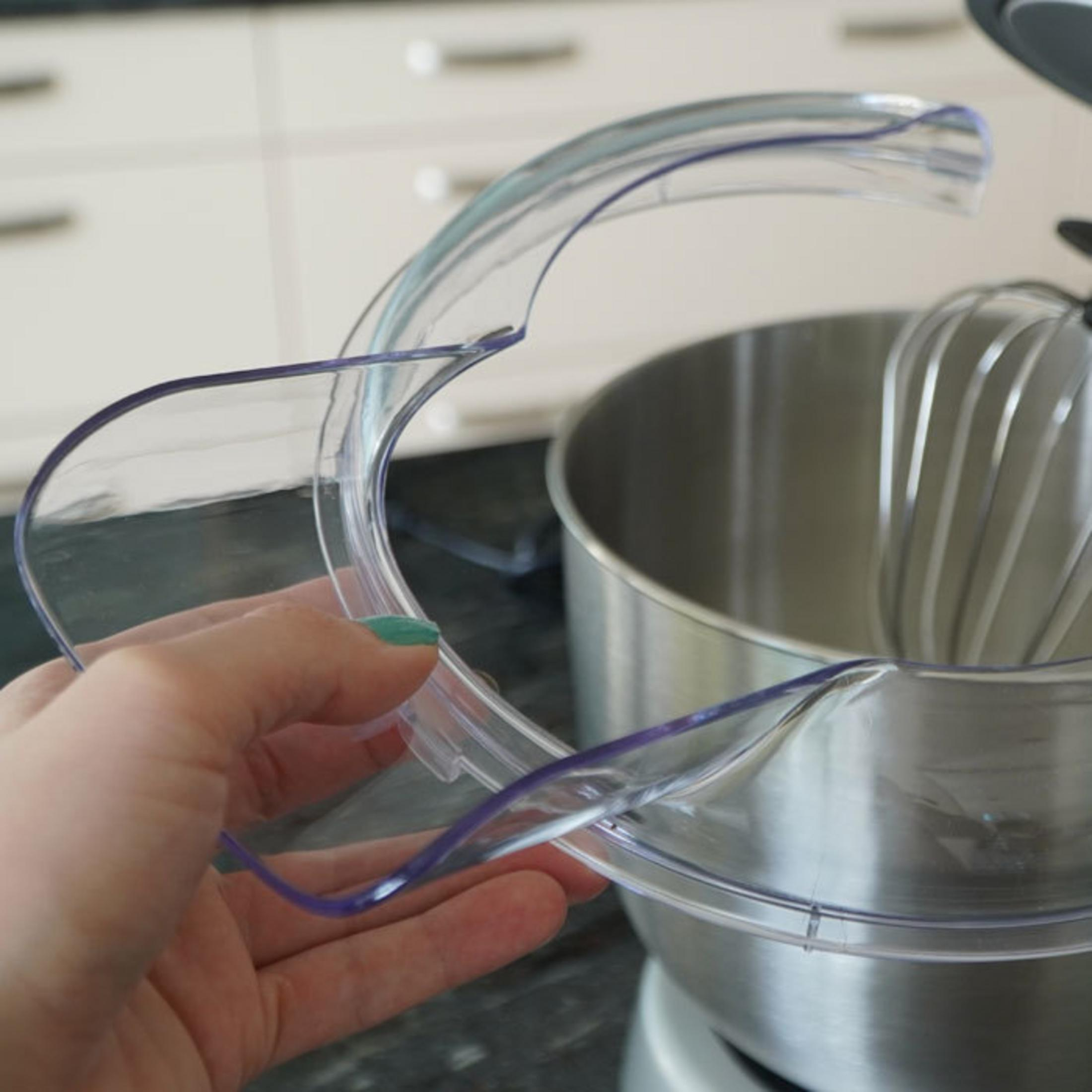 Una mano tiene un paraspruzzi trasparente sopra un robot da cucina con ciotola in metallo.