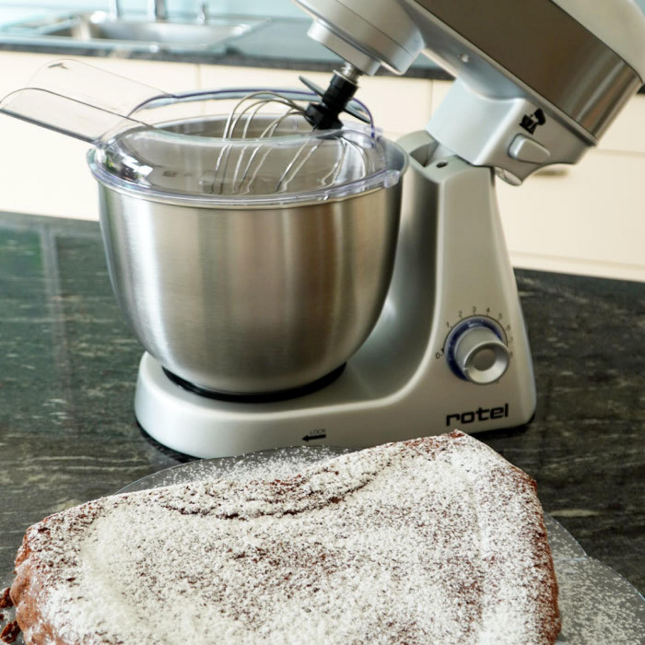 Robot da cucina argentato con ciotola e frusta, accanto una torta spolverata di zucchero a velo.