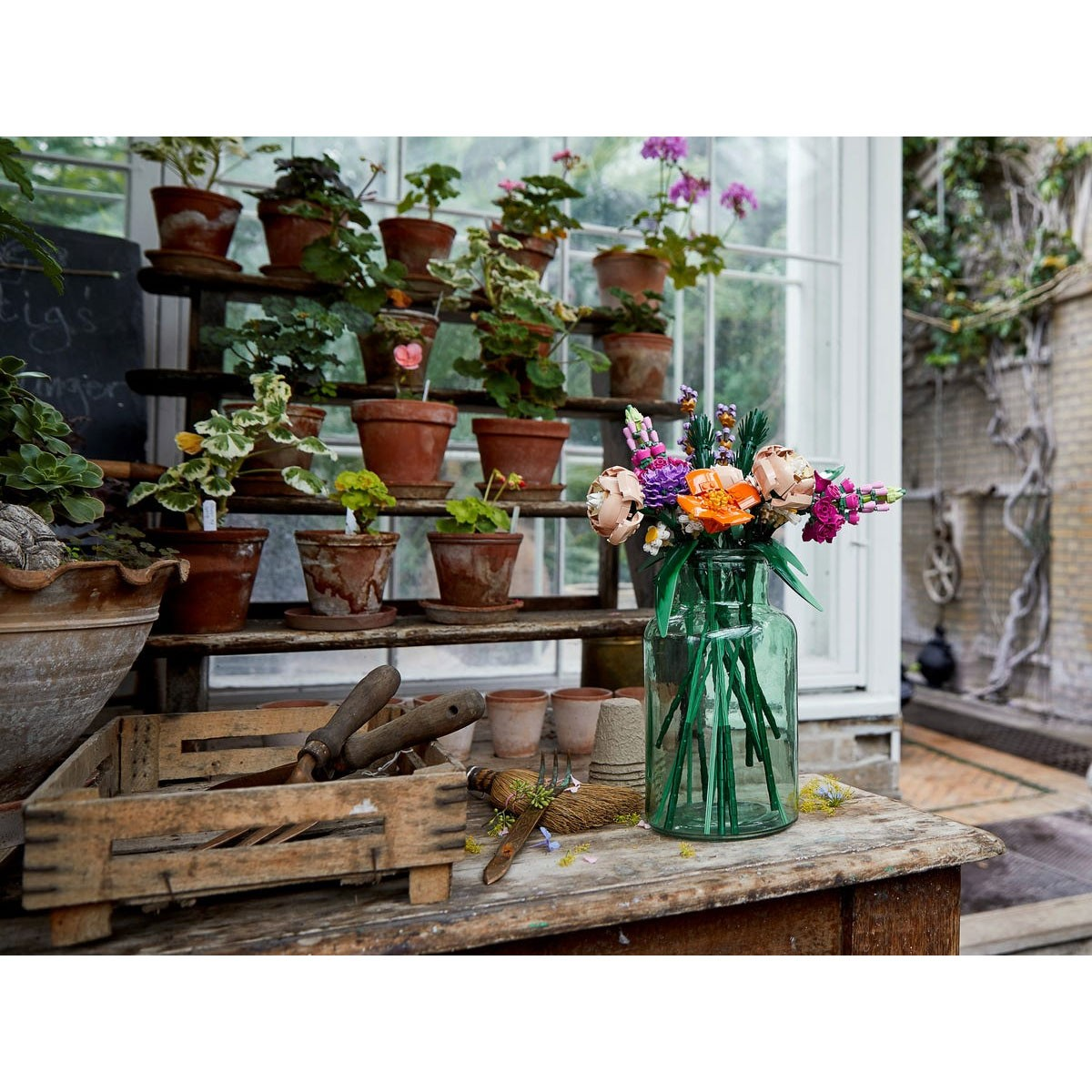 Flores en un jarrón verde sobre una mesa de madera con plantas al fondo.