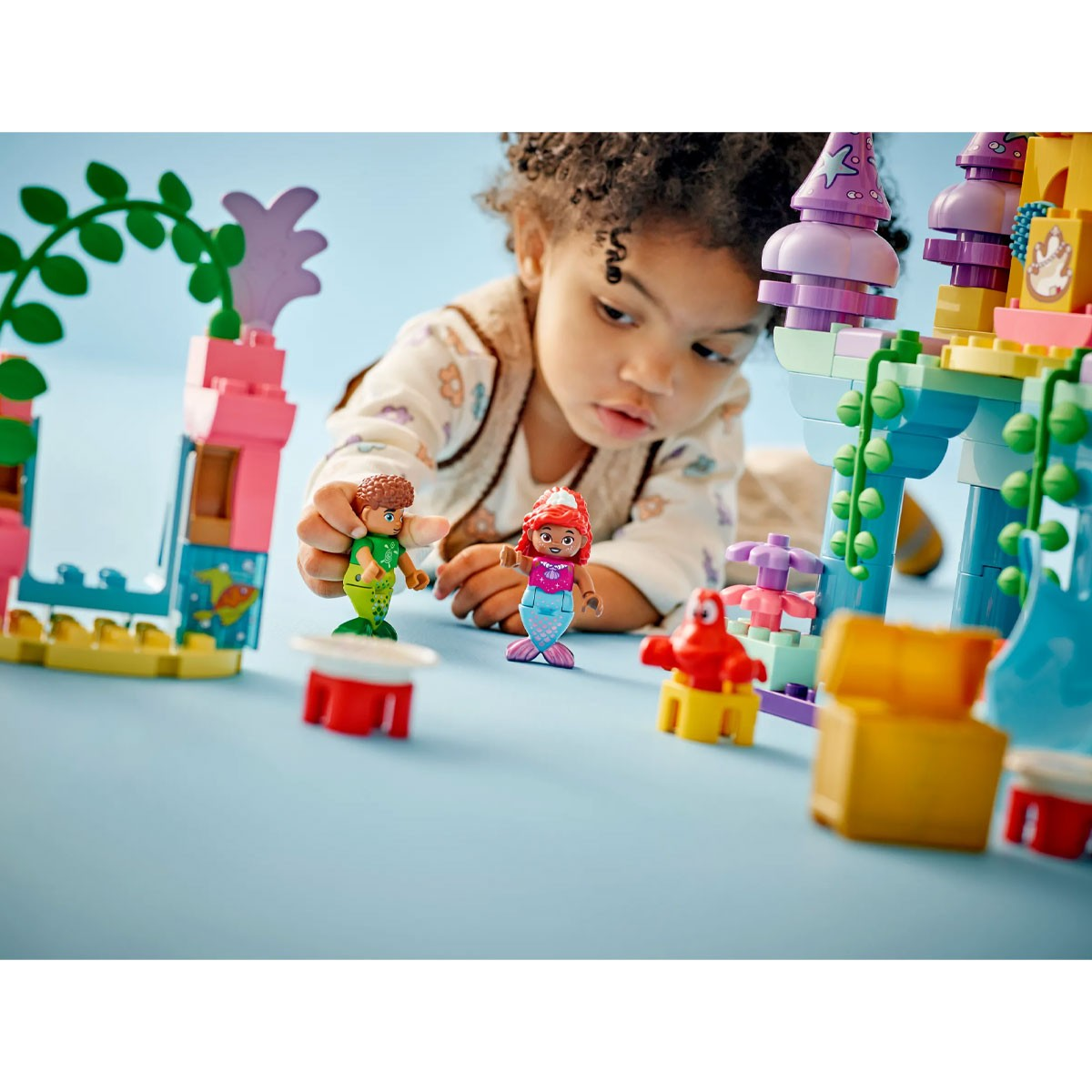Dziecko bawi się figurkami syrenek Lego i zamkiem na niebieskim tle.