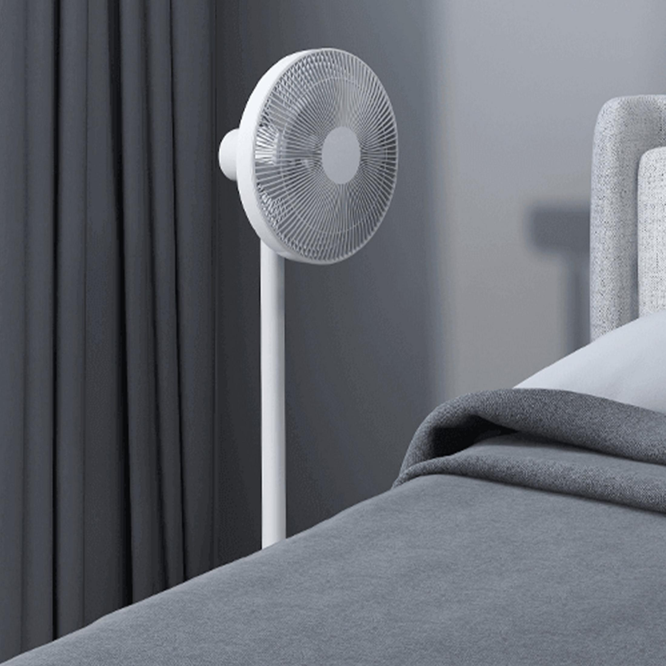 Biały wentylator stojący stoi obok łóżka z szarą pościelą.